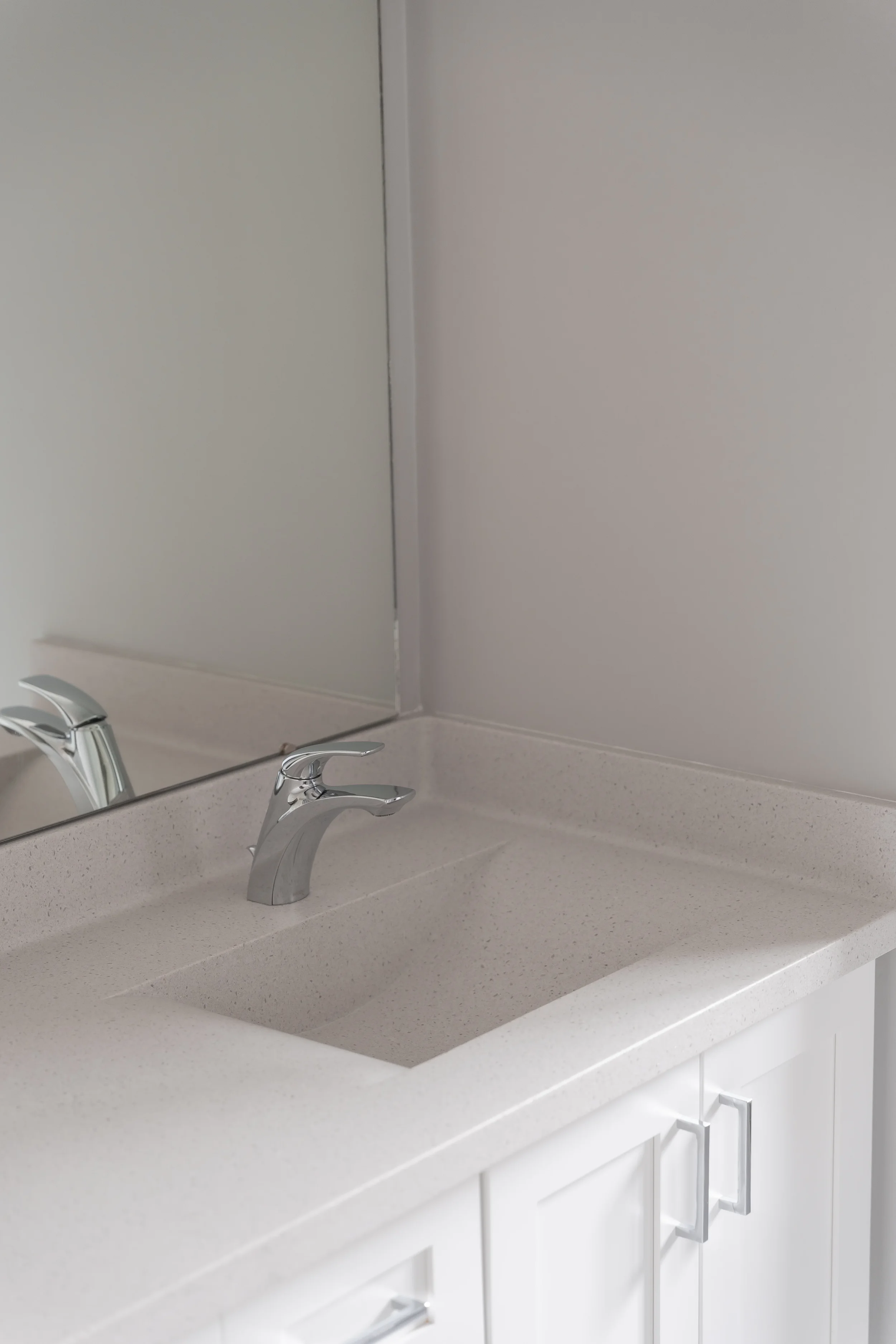 White bathroom vanity with dual stainless steel faucets, beige speckled countertop, large mirror, and white cabinets with silver handles.