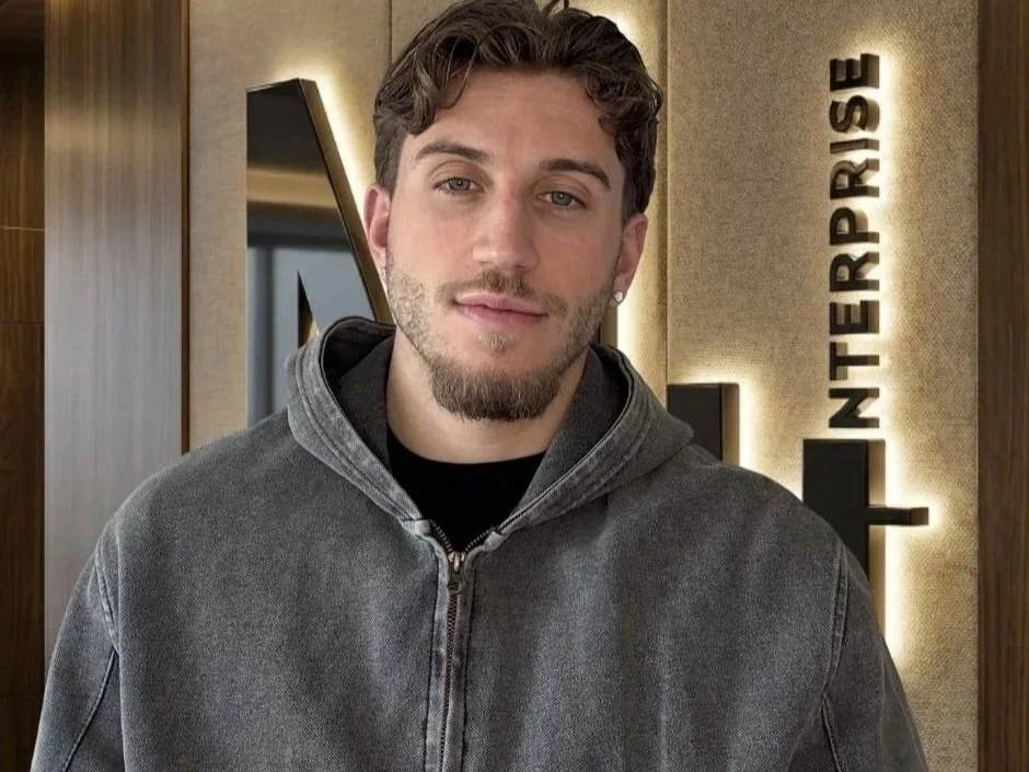 Ein junger Mann mit braunen Haaren und einem schwarzen T-Shirt, trägt eine graue Zip-Jacke, steht vor einer Wand mit dem Schriftzug 'INTERPRESS' in einem modernen Büro oder Empfangsbereich.