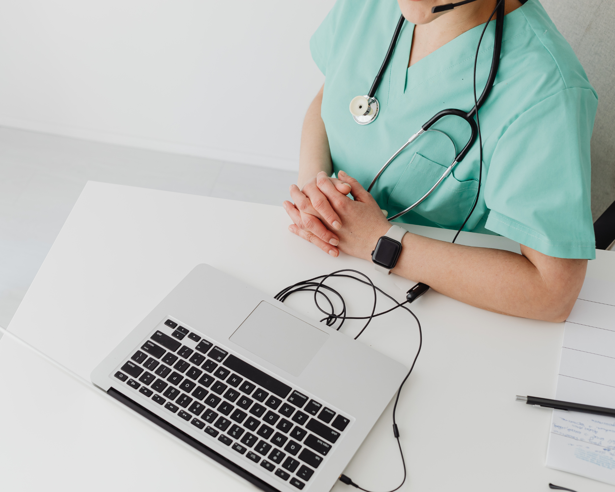 Ärztin in medizinischer Kleidung mit Stethoskop und Smartwatch sitzt an weissem Tisch mit Laptop und Notizblock.