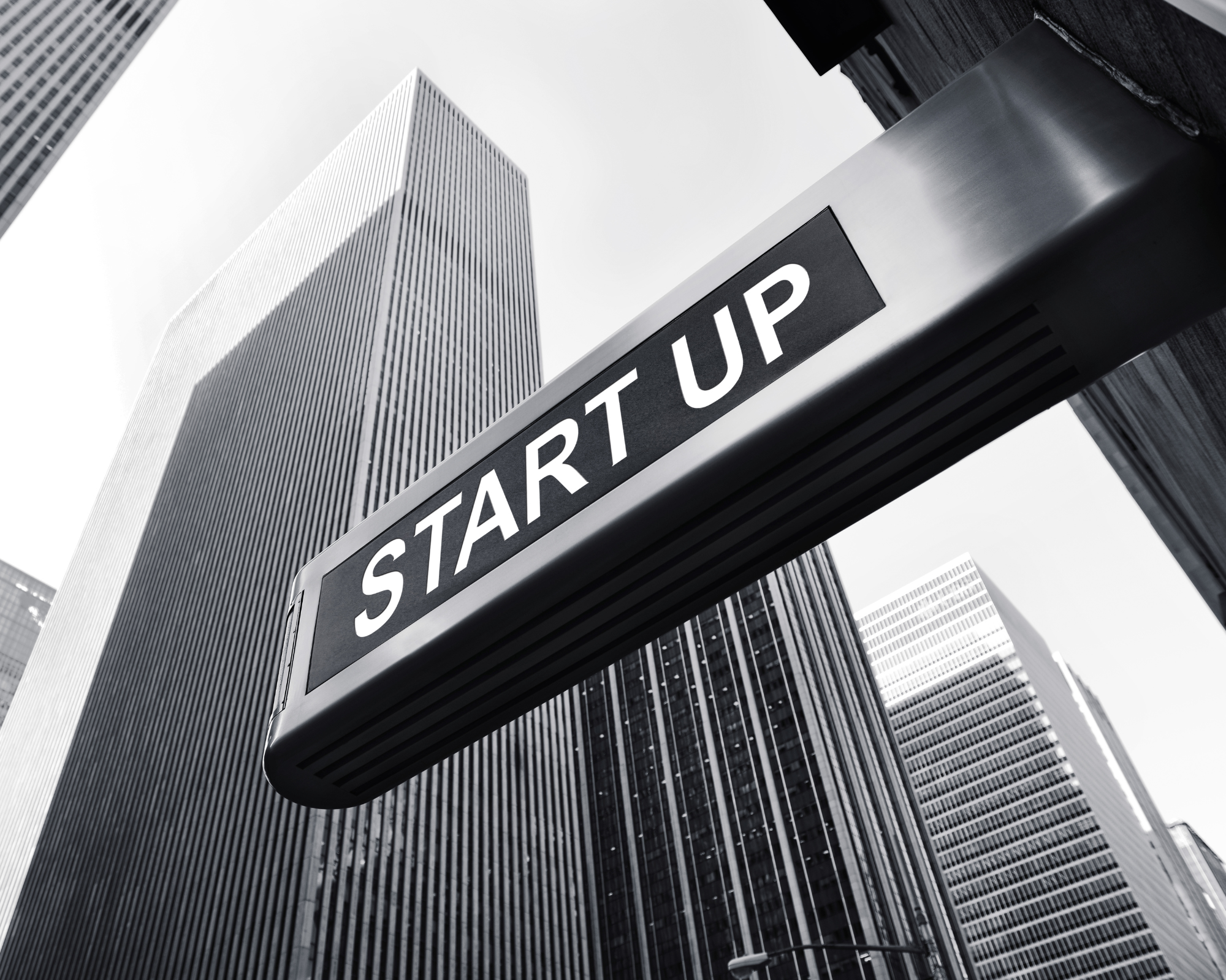 Ein Start-up-Schild vor Hochhäusern in einer Stadt, schwarz-weiss, perspektivischer Blick nach oben.