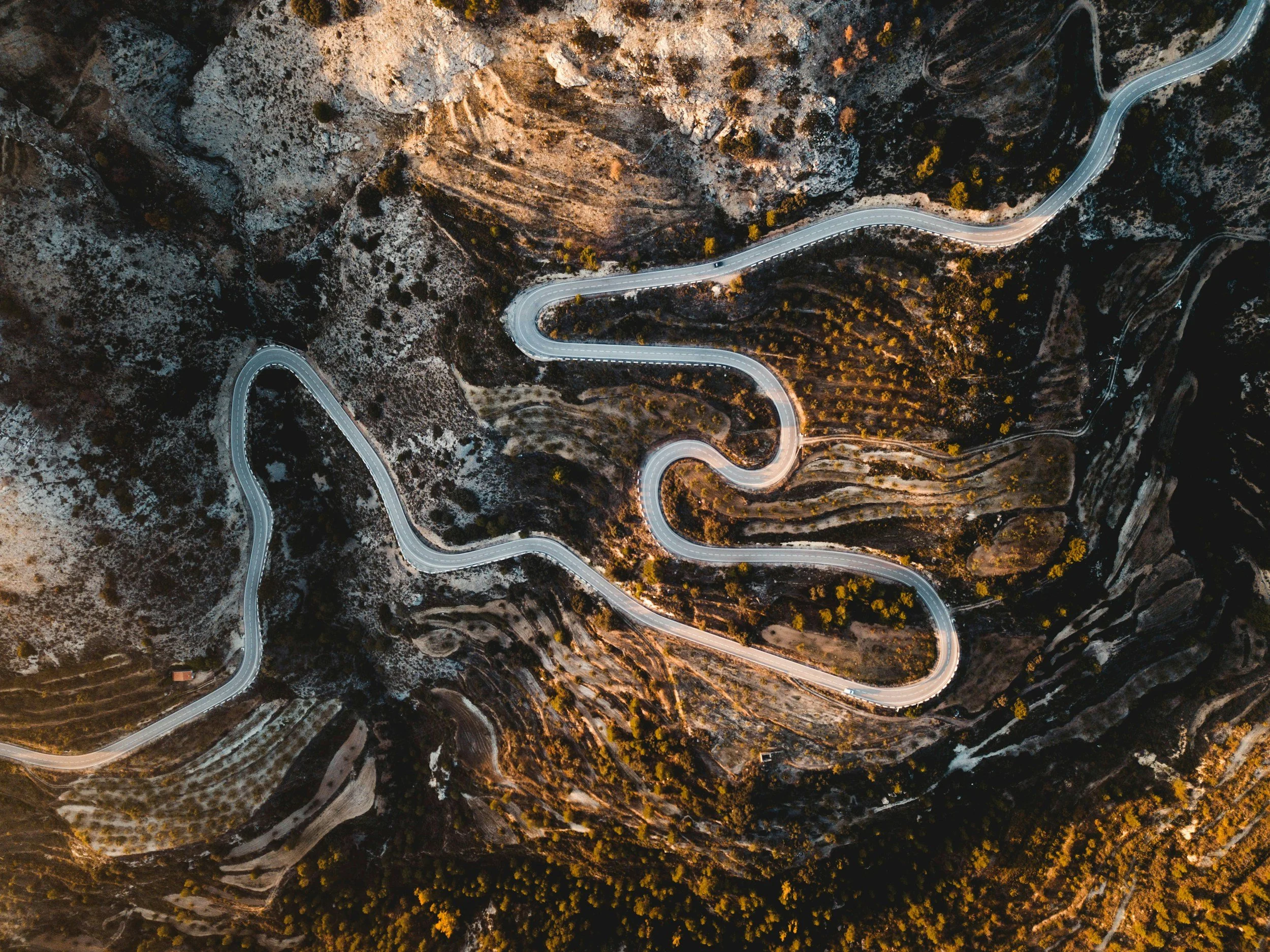 An aerial view of a winding mountain road through a rugged, desert landscape with sparse vegetation and rocky terrain.
