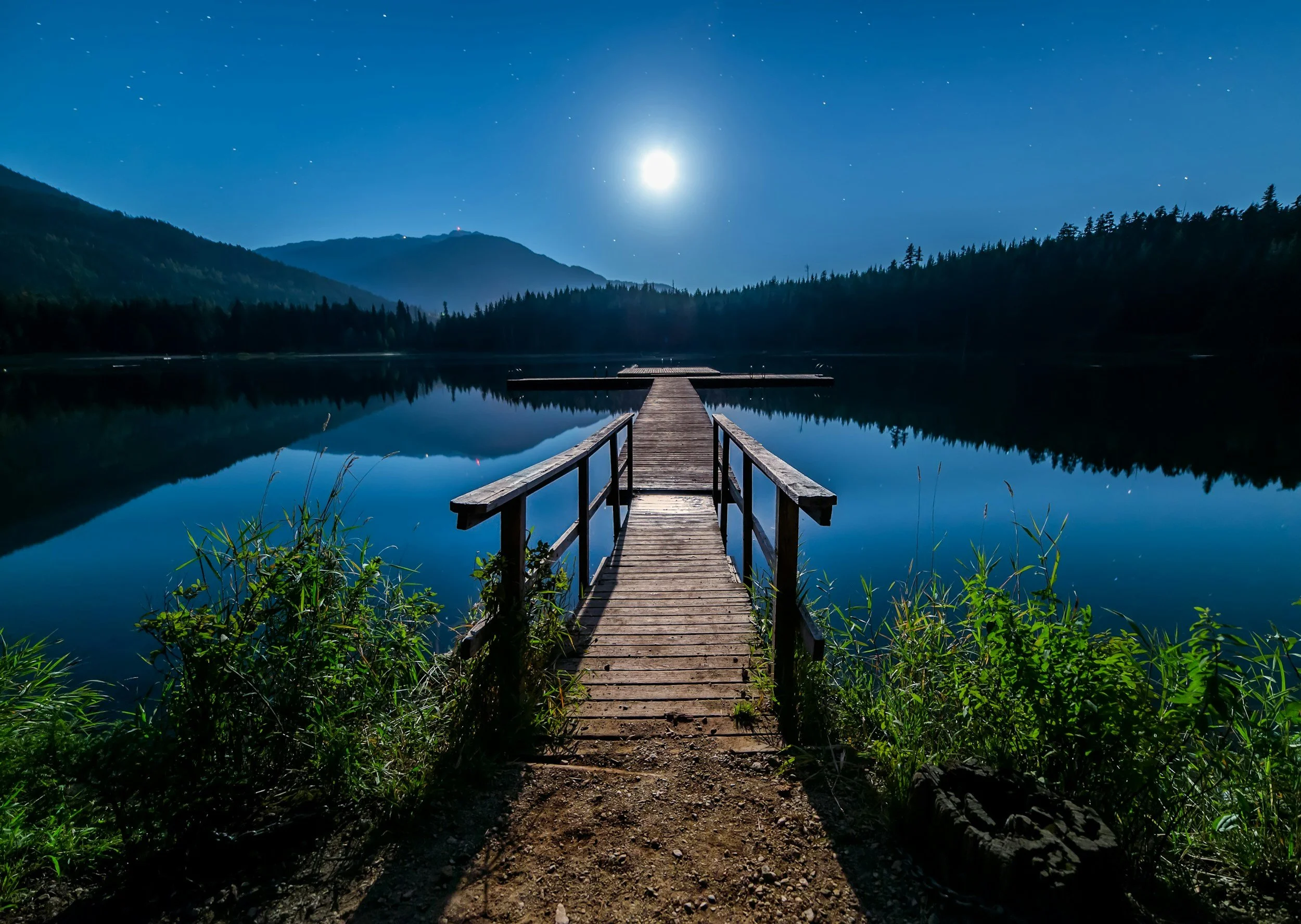 Nighttime scene of a wooden dock extending into a calm lake, illuminated by a bright full moon, surrounded by forested mountains, with clear reflections in the water and a starry sky overhead.