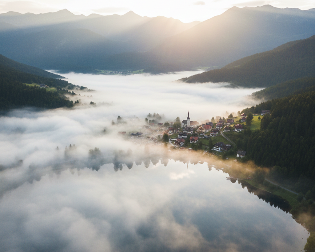 A foggy valley with a small village, a church with a tall steeple, surrounded by forested hills and mountains at sunrise.