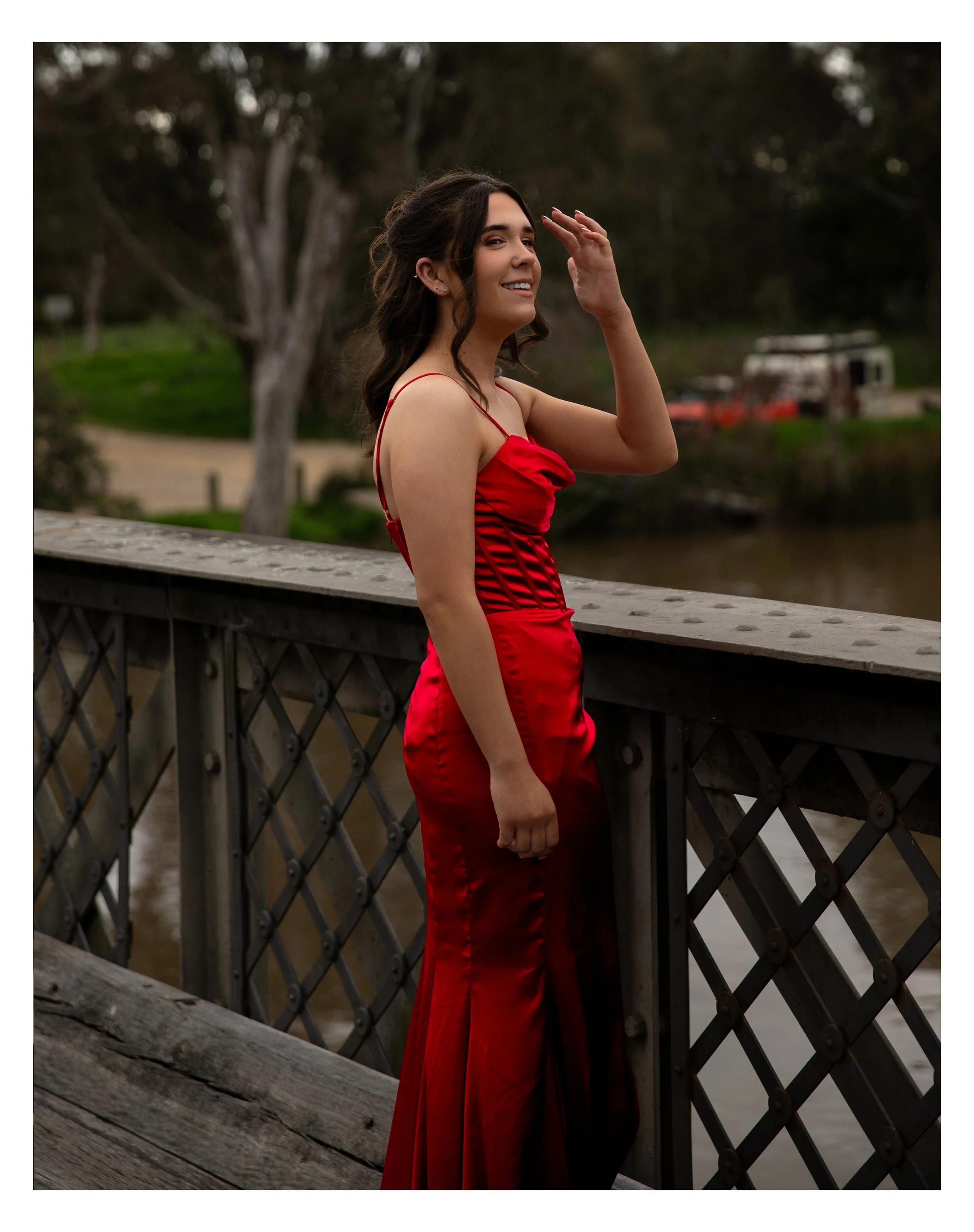 A woman in a red evening gown stands on a bridge by a river, smiling and waving her hand.