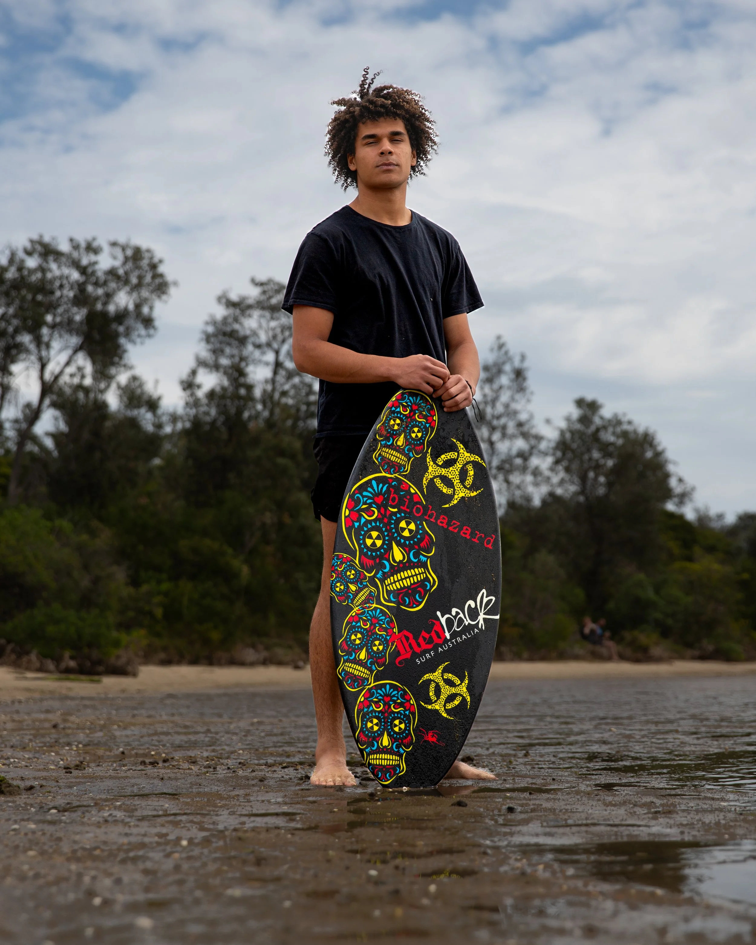 A young man standing barefoot on a beach holding a colorful skateboard with skull and biohazard designs, wearing a black T-shirt, with trees and cloudy sky in the background.