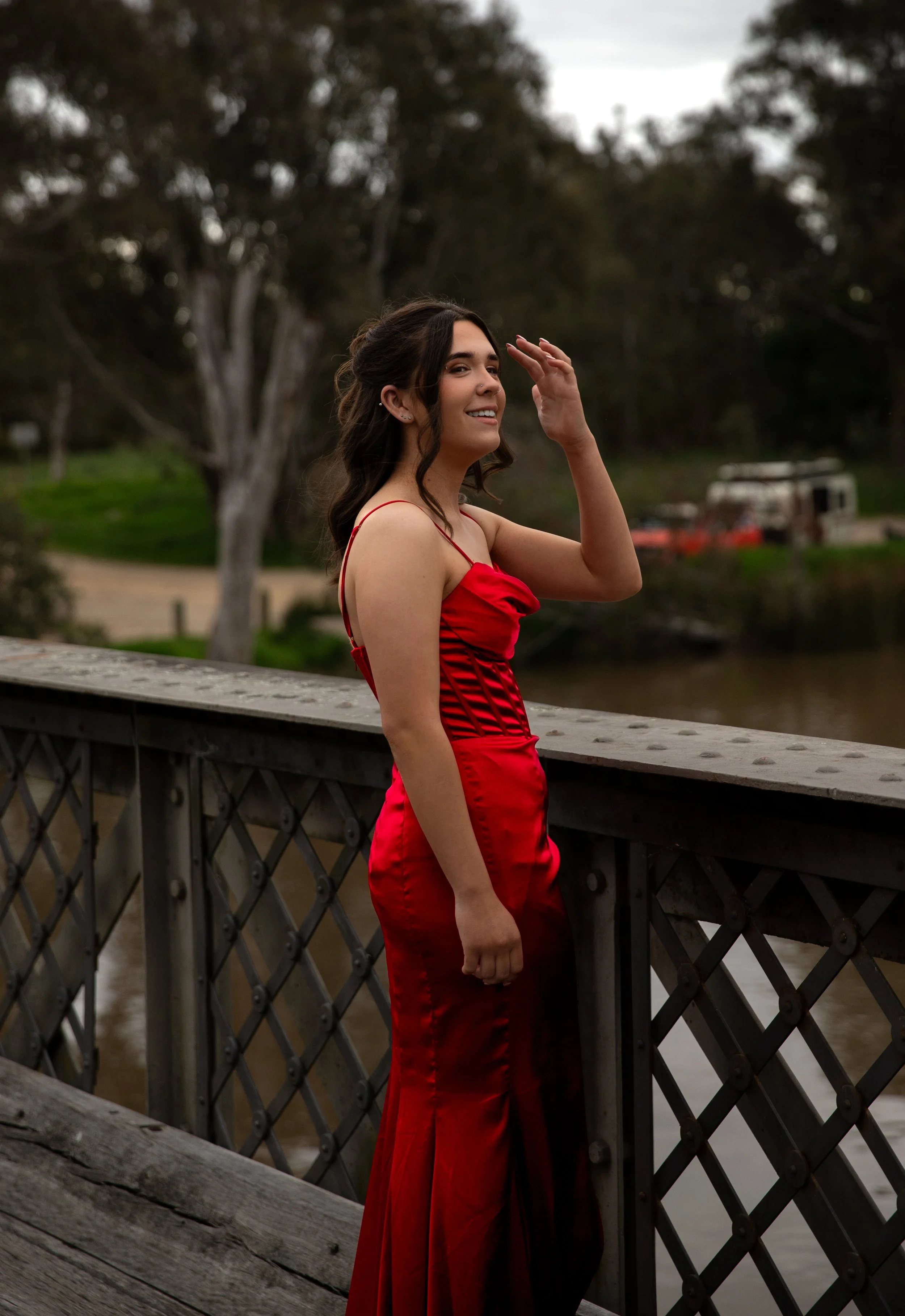 A young woman in a red satin dress standing on a bridge near a body of water, with trees and a vehicle in the background.