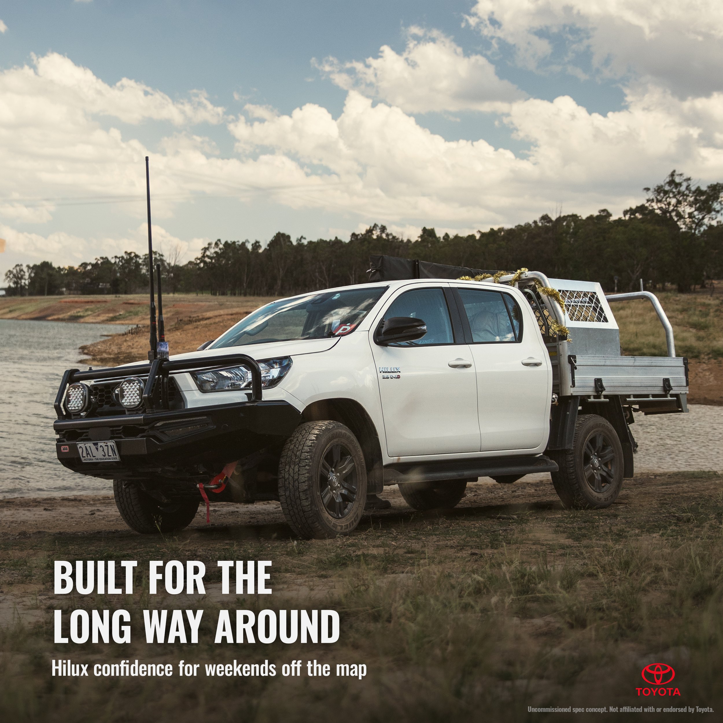 A white Toyota Hilux pickup truck parked near a river, with a rural landscape and trees in the background. The truck has a black bull bar, mounted lights, and antennas, indicating it is built for off-road adventures.