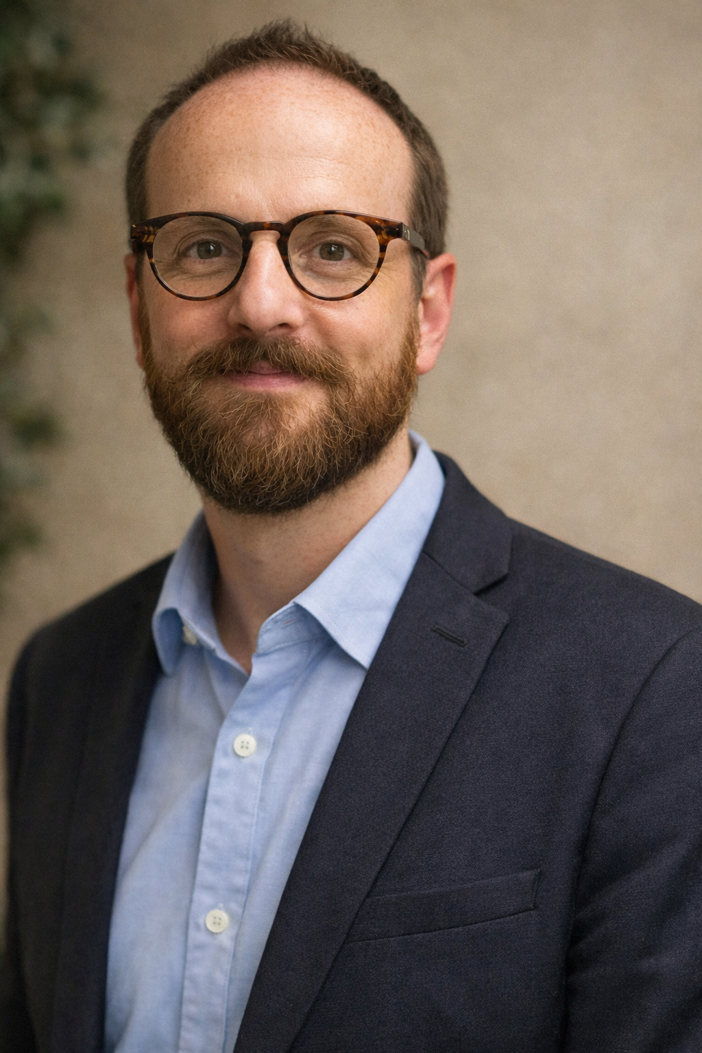 A man with glasses, a beard, and short hair, wearing a dark suit jacket over a light blue shirt, standing against a plain background.