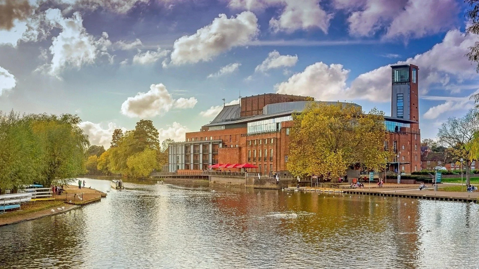 Scene of a river with trees on both sides, a modern building with red brick and glass windows, and a blue sky with fluffy white clouds.