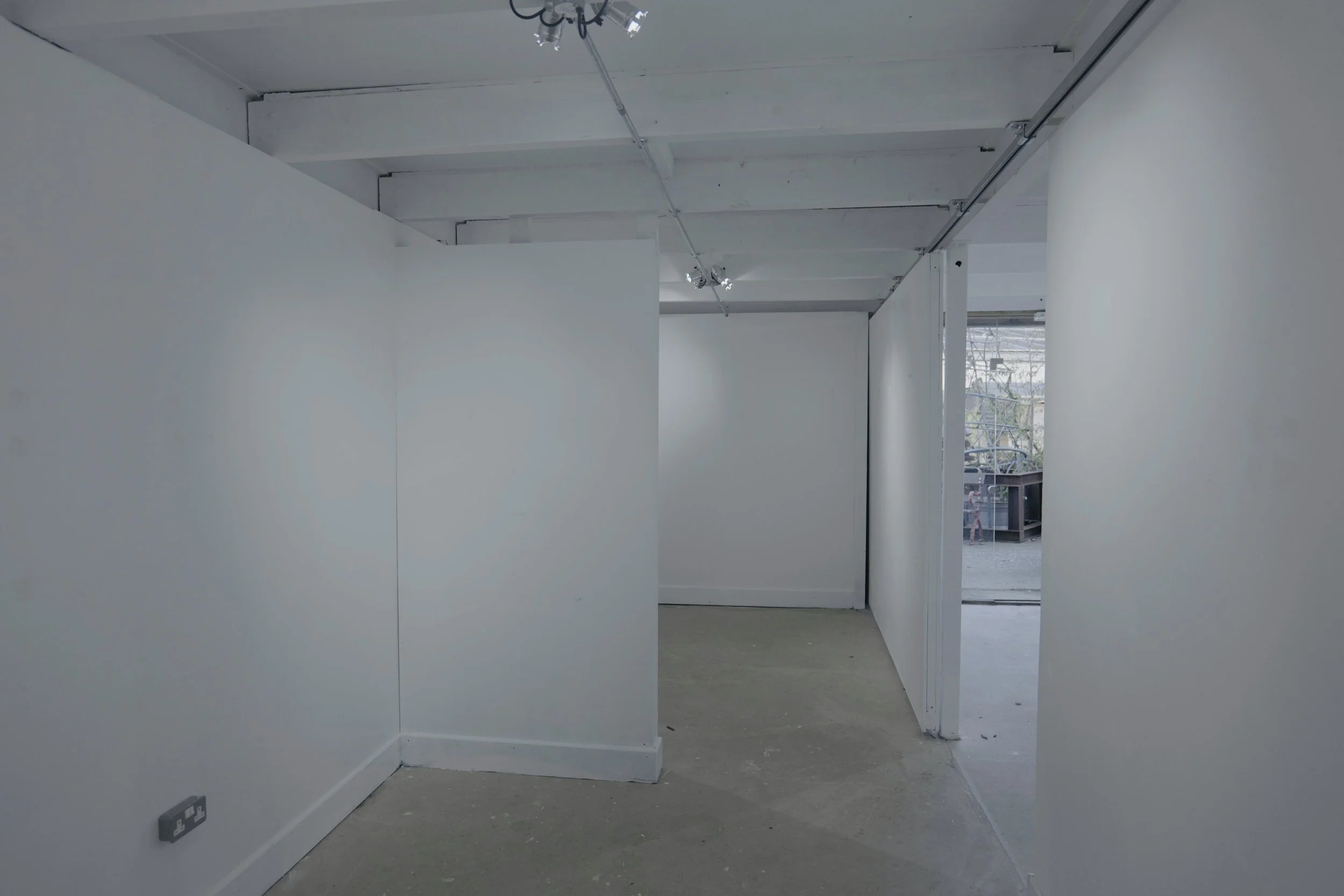 Empty neighboring walls in a white-walled room with electrical outlet on the lower left wall and visible ceiling track lighting, with an open doorway leading to outside area.