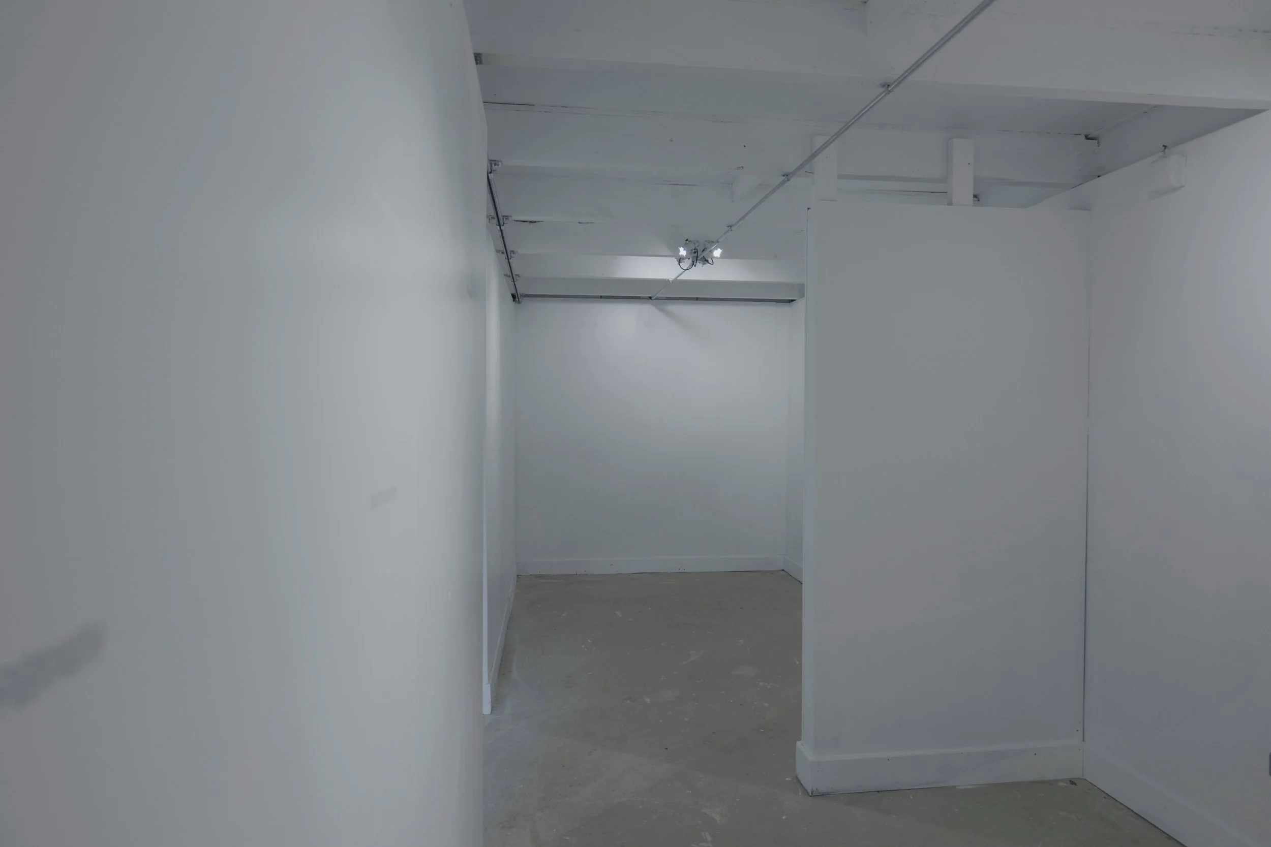Empty white-walled gallery space with overhead lighting and unfinished concrete floor.