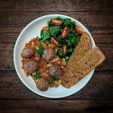 White plate containing meatball and pasta dish, mixed greens salad with cherry tomatoes and pomegranate seeds, and two slices of bread, on a wooden table.