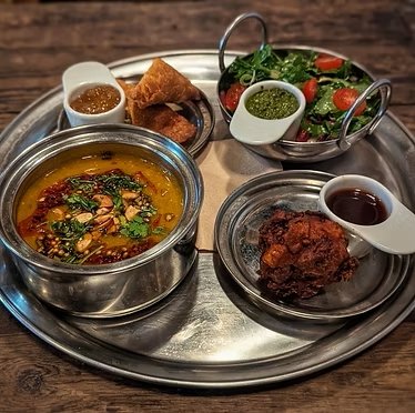 Indian meal with side dishes on a metal tray, including lentil soup, fried appetizers, salad, and a curry with sauce.