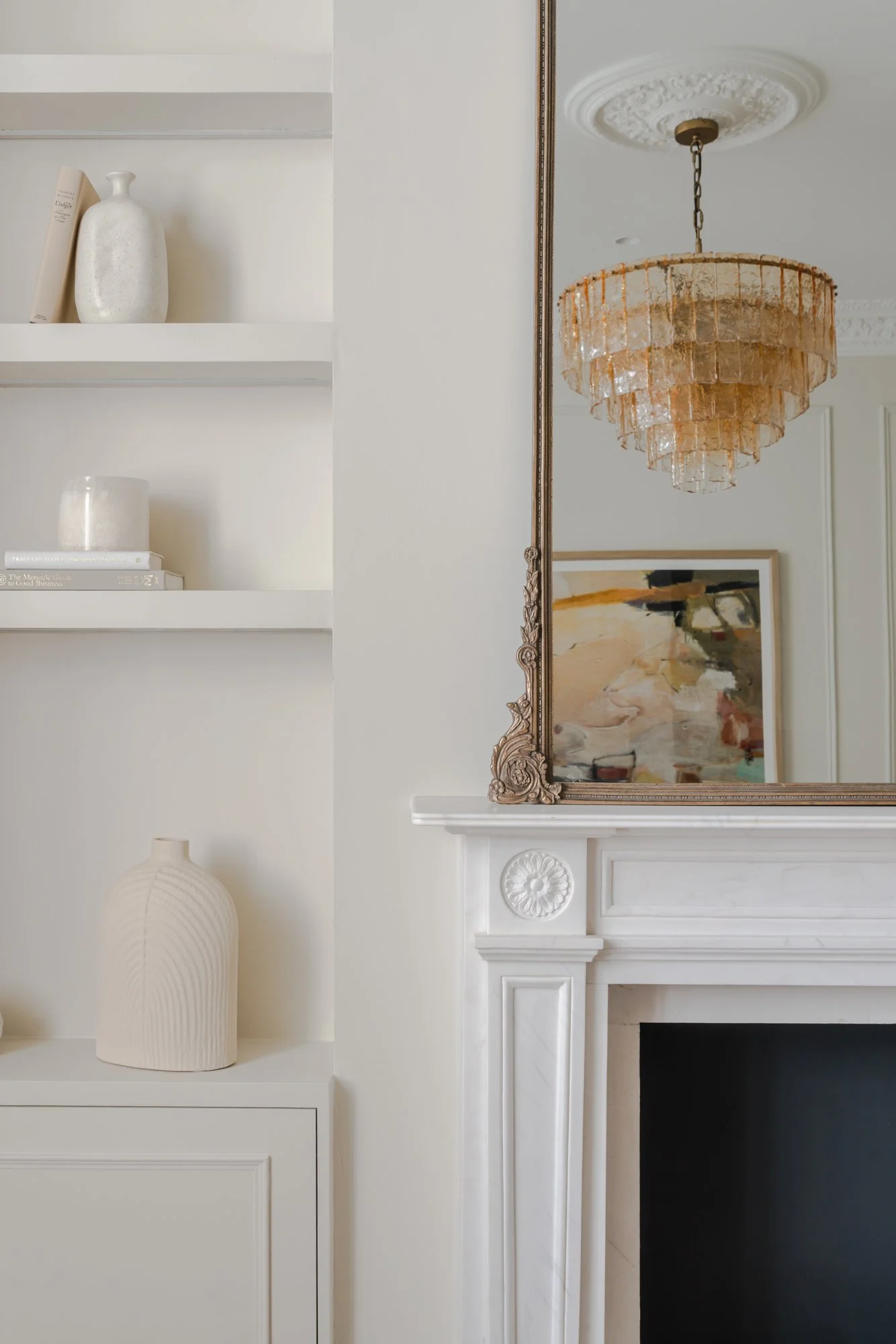 reflection in a mirror showing a chandelier, painting, and part of a white fireplace mantel with decorative molding, and built-in shelves with vases and books.