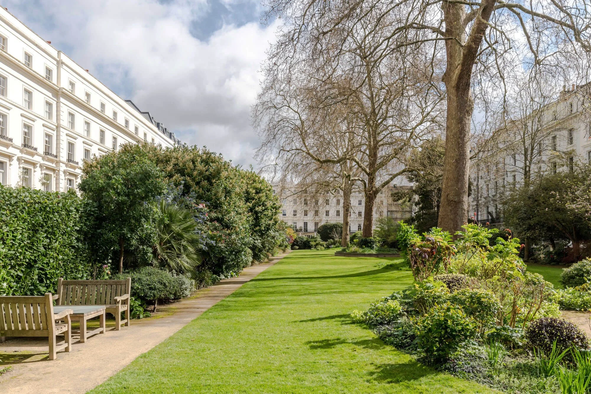 A peaceful city park with trimmed grass, bushes, and tall trees, surrounded by white residential buildings, with a cloudy sky overhead.