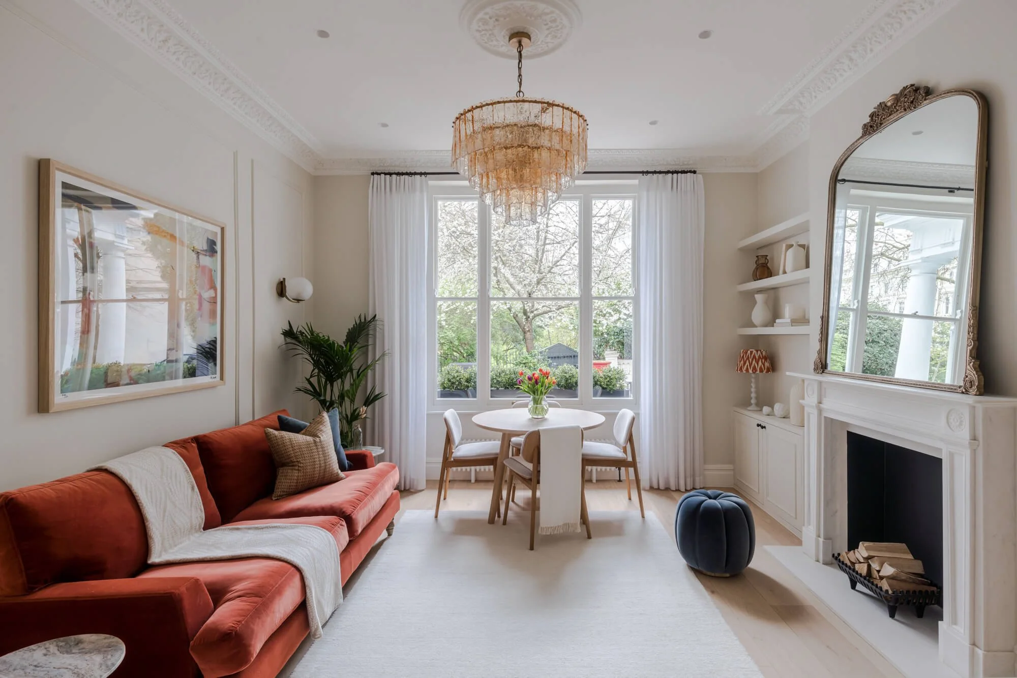 Bright living room with a large window, white curtains, beige walls, a red velvet sofa with throw pillows, a round white dining table with four chairs, a decorative chandelier, a fireplace with a mirror above, and built-in shelves with decor items.