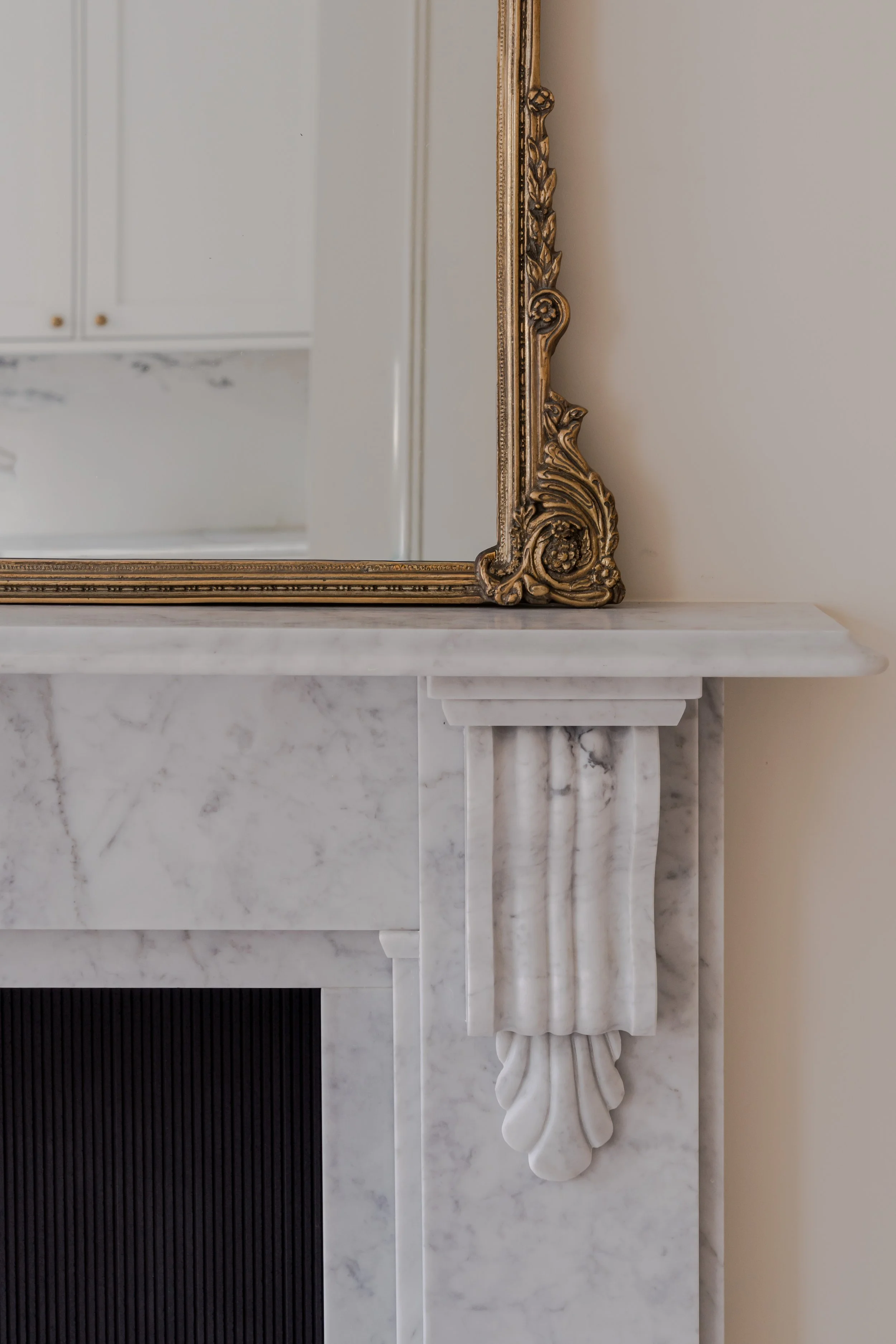 Close-up of a marble fireplace mantel with ornate corbel detail, a large ornate gold-framed mirror on top, and a partial view of a kitchen in the background.