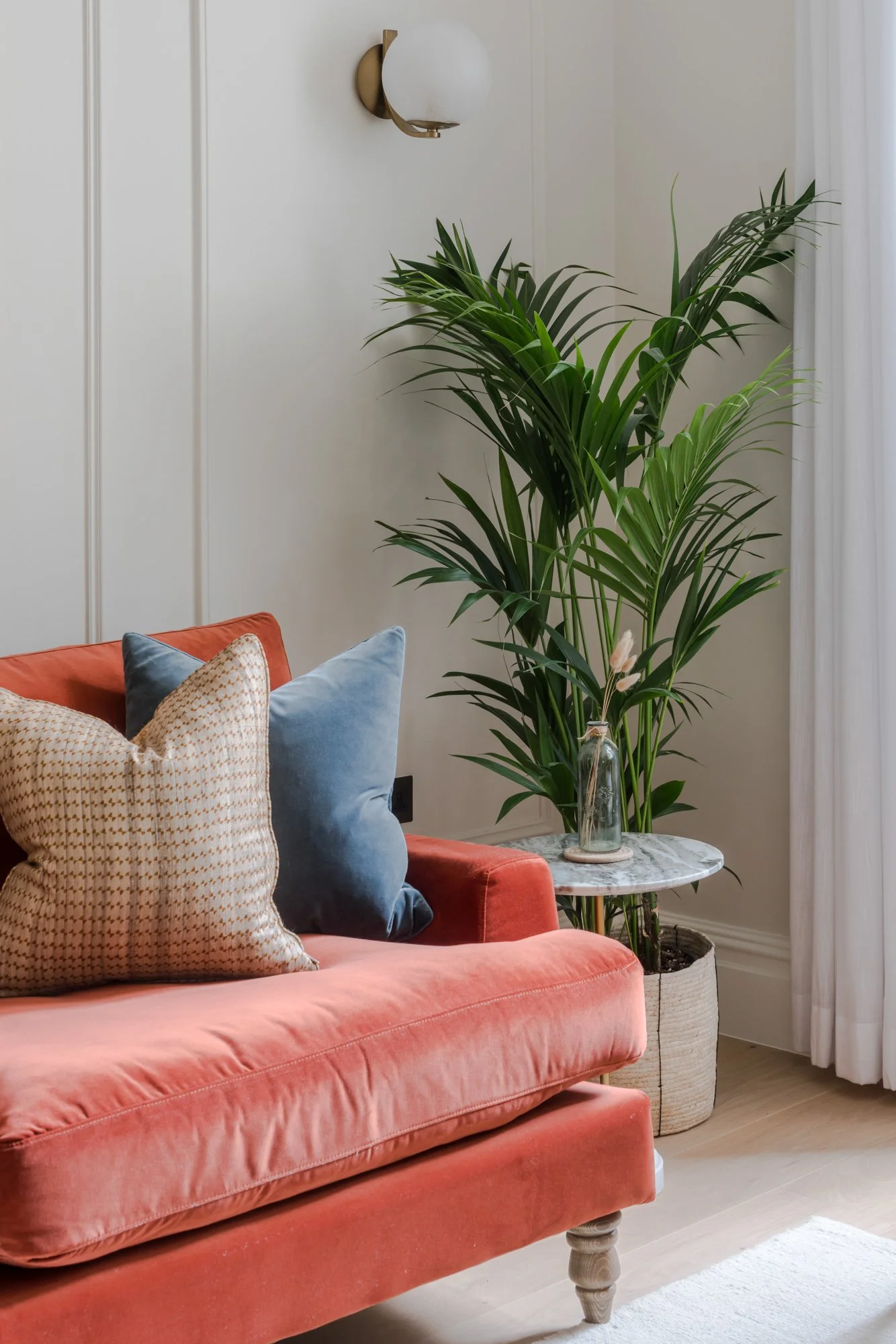 A cozy living room corner featuring a coral velvet sofa decorated with textured and velvet pillows, a large green potted plant next to a small round marble-topped side table with a clear glass vase holding dried flowers, all illuminated by a wall-mounted modern light fixture.