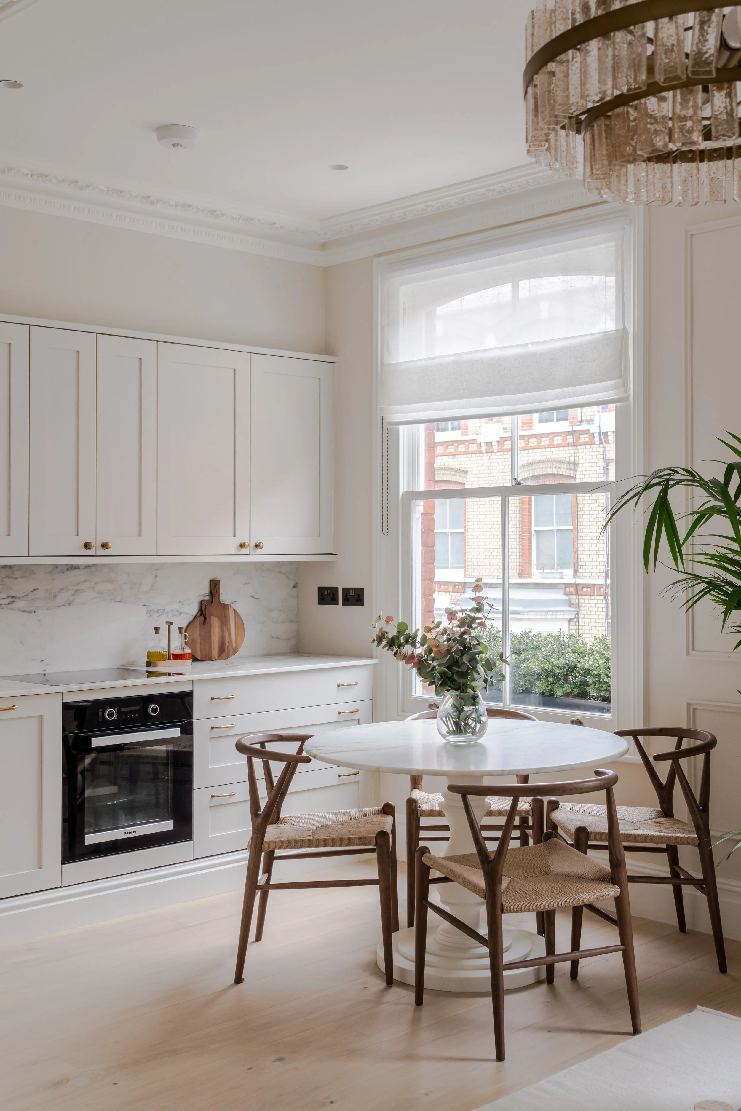 Bright kitchen with white cabinets, marble backsplash, and a round dining table with four wooden chairs. A large window lets in natural light, decorated with two shades, and there is a vase of flowers on the table and a potted plant nearby.