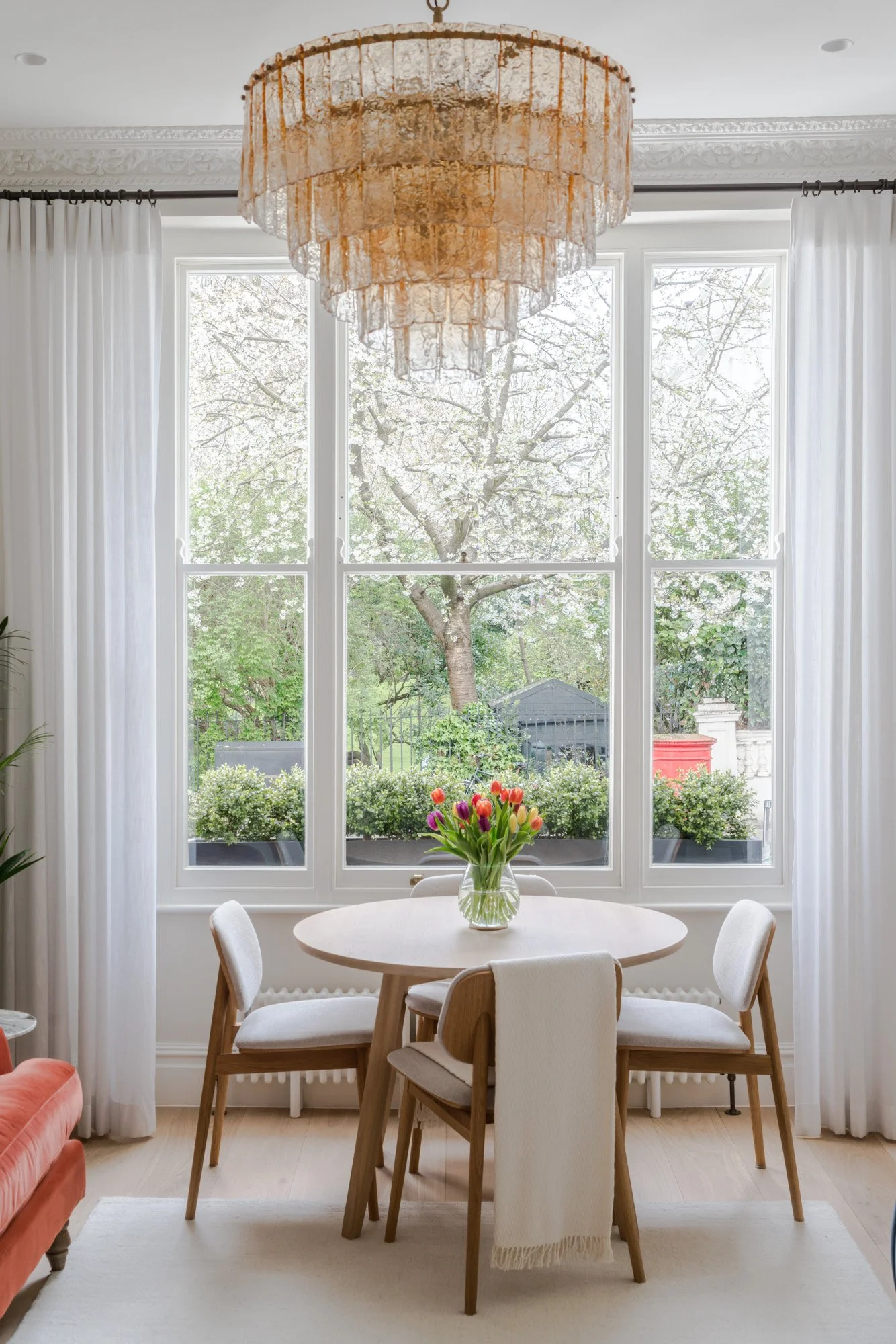 A dining room with a round table, four chairs, and a vase of colorful tulips, set in front of large windows with white curtains, overlooking a garden with blooming trees.