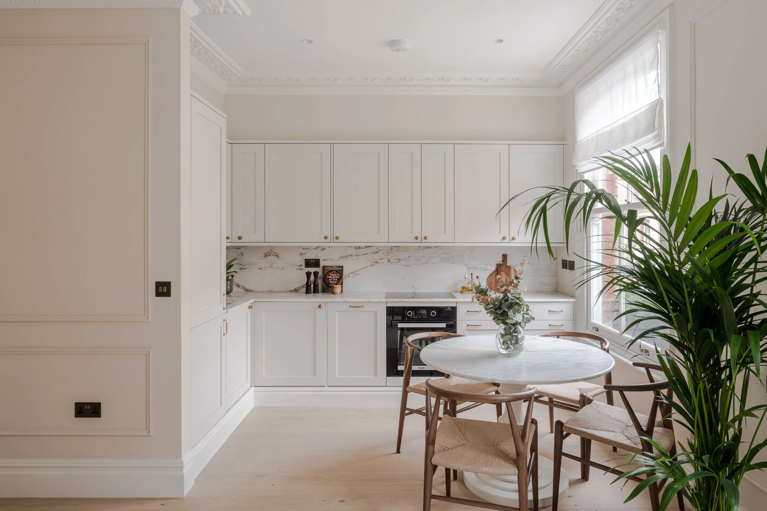 A bright kitchen with white cabinets, a marble backsplash, and a dining area with a round table and four wooden chairs, decorated with a vase of flowers, green plants, and some kitchen accessories.