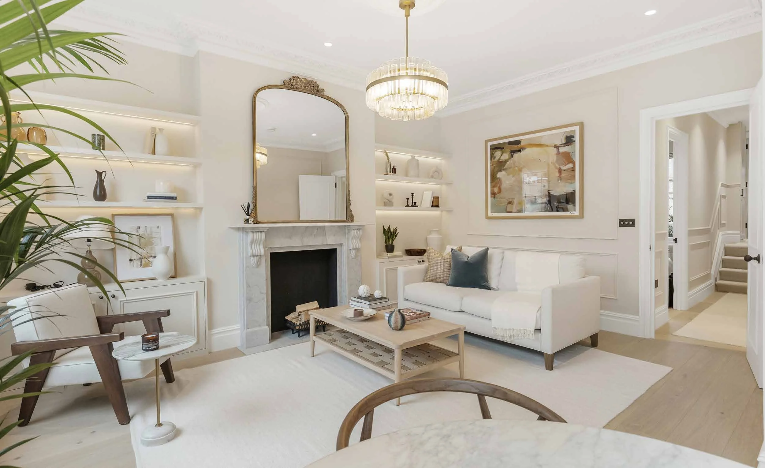 Bright living room with white walls, large windows, and elegant fireplace with mirror. Decor includes houseplants, books, and neutral-toned furniture.