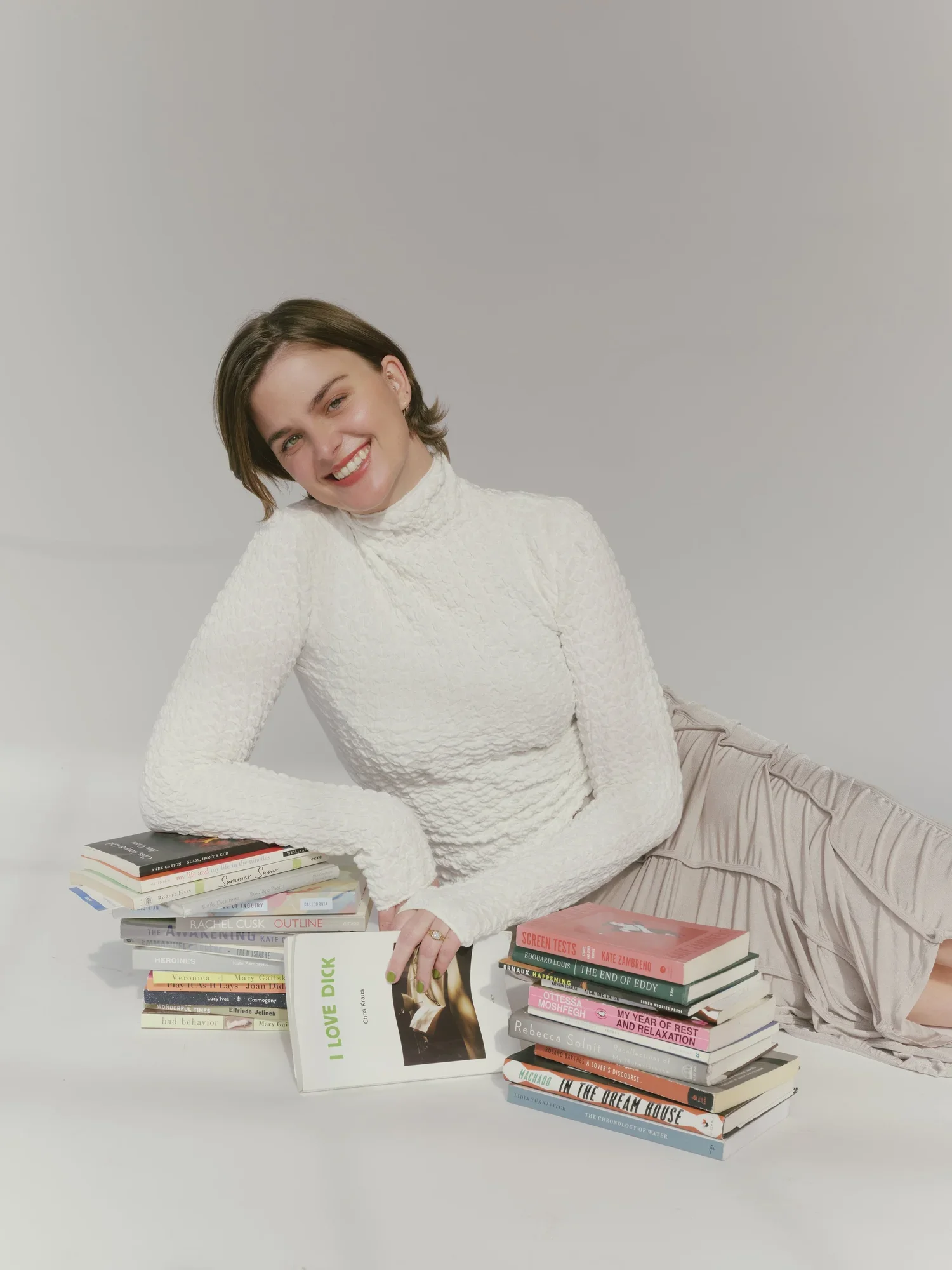 Photo of Ashley Pattison Scott, A woman with short brown hair and light skin, wearing a white textured turtleneck top, smiling and leaning on a stack of books, with another pile of books in front of her, on a white background.