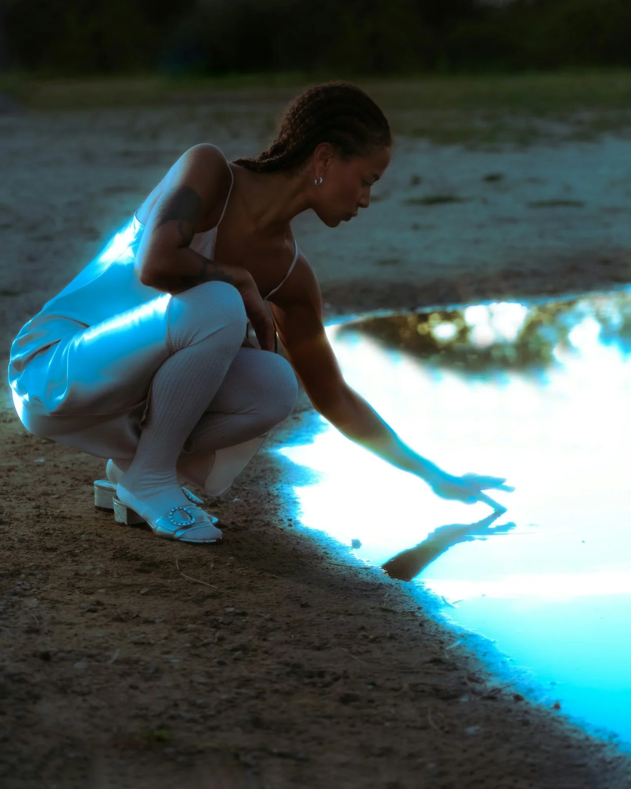 A woman dressed in white squat by a lake at dusk, touching the water with her hand. She has braided hair and tattoos on her arm.