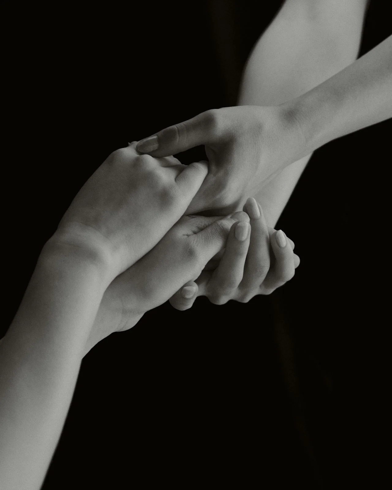 Two hands gently holding each other, one hand with fingers intertwined with the other, against a dark background.