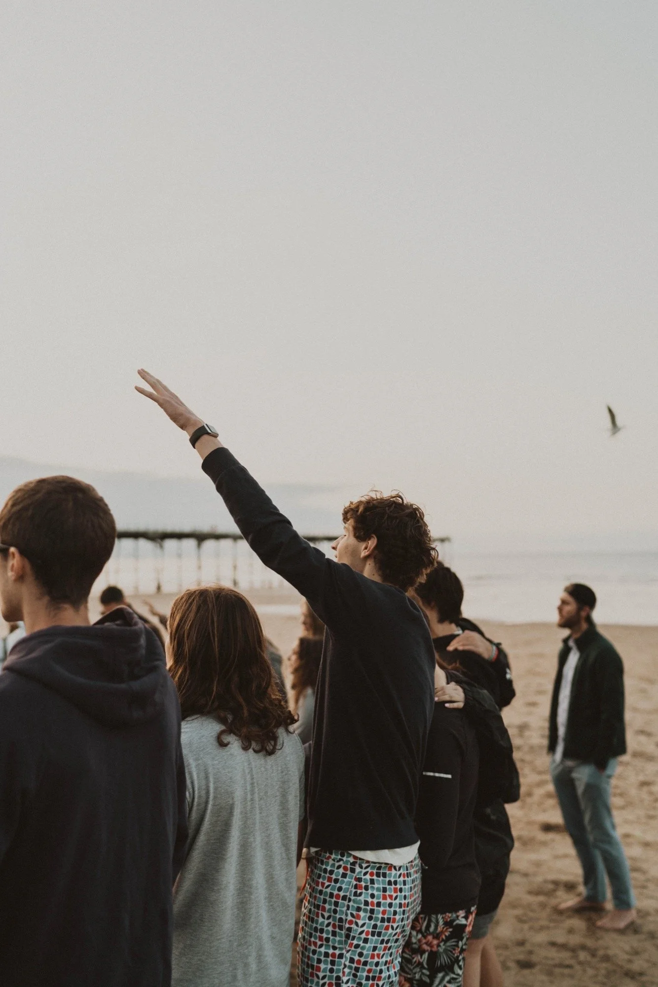 Group of people standing on a beach, with one person raising his hand, and the ocean and pier in the background.