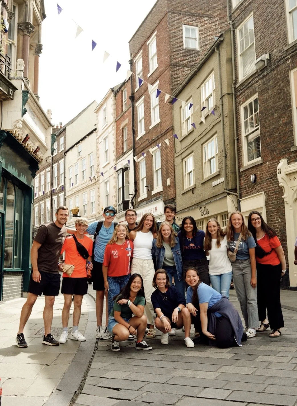Group of 14 young people smiling and posing on a city street with brick and historic buildings, some with storefronts, and purple and white bunting overhead.