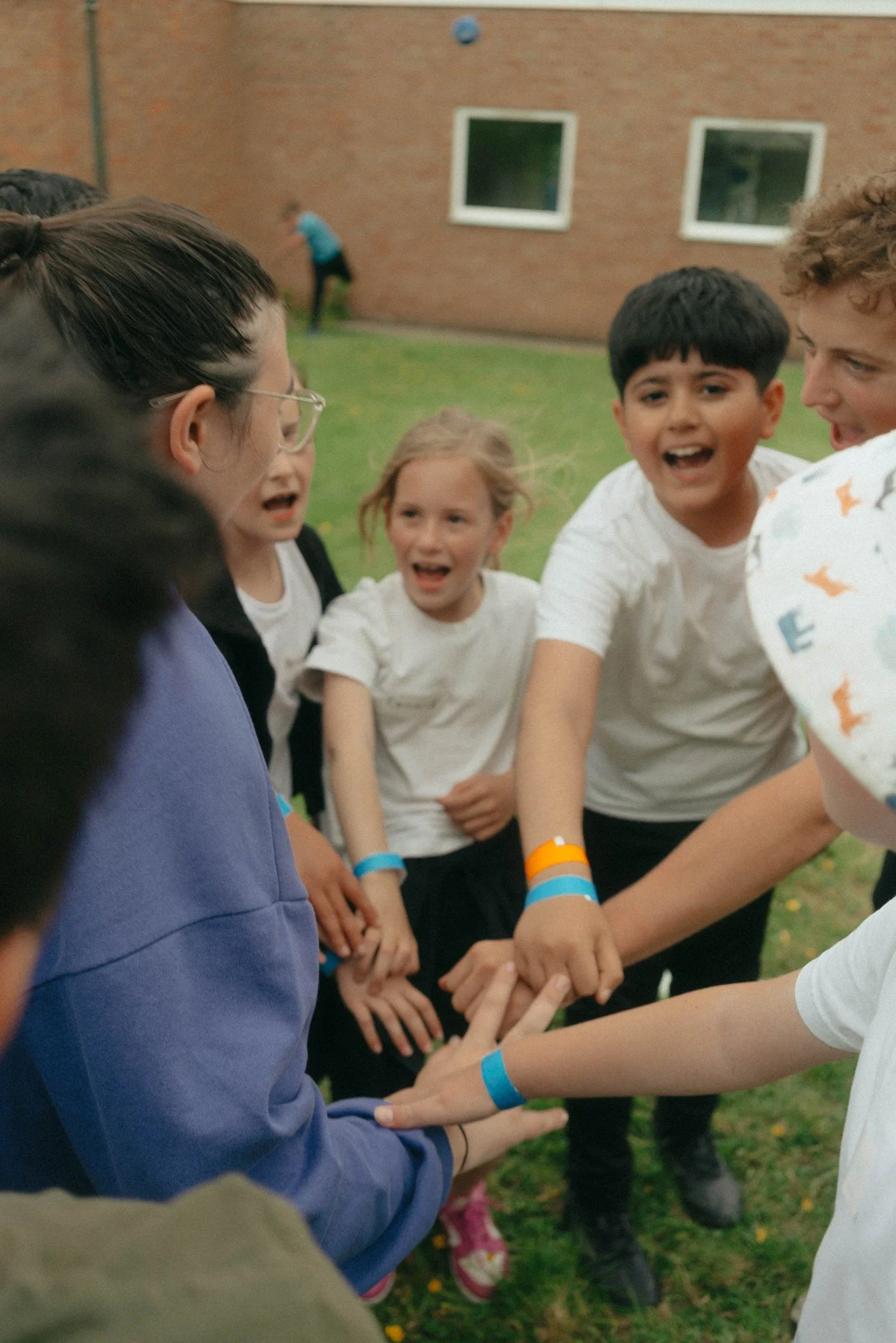 Group of children and a woman holding hands in a circle outdoors, engaging in a team activity on grass near a brick building.