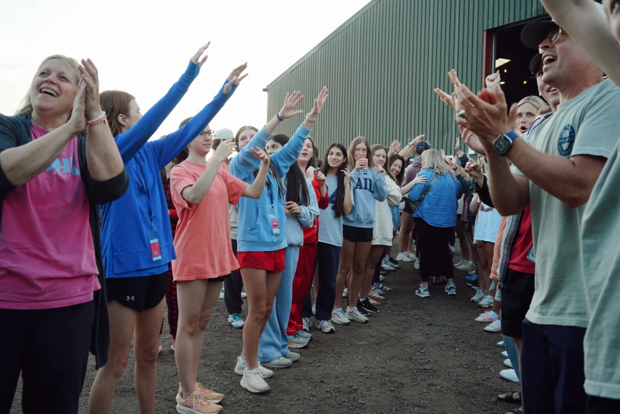 Group of people, including women and children, standing in two lines facing each other outdoors near a green building, clapping and cheering.