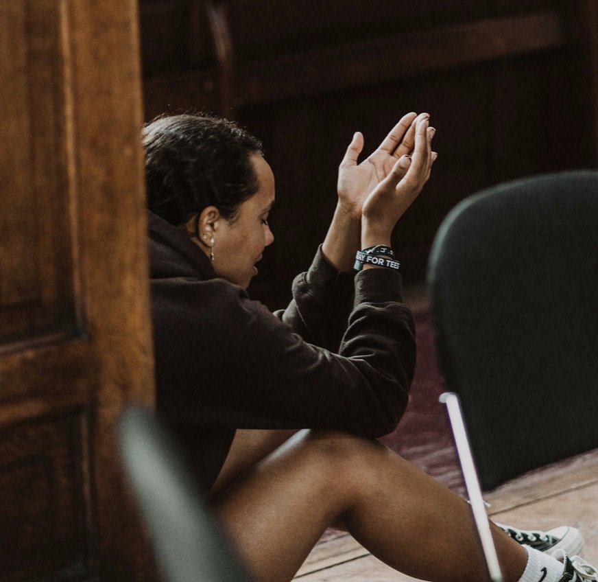 A woman sitting on the floor near a wooden wall, with her hands raised in prayer or contemplation.