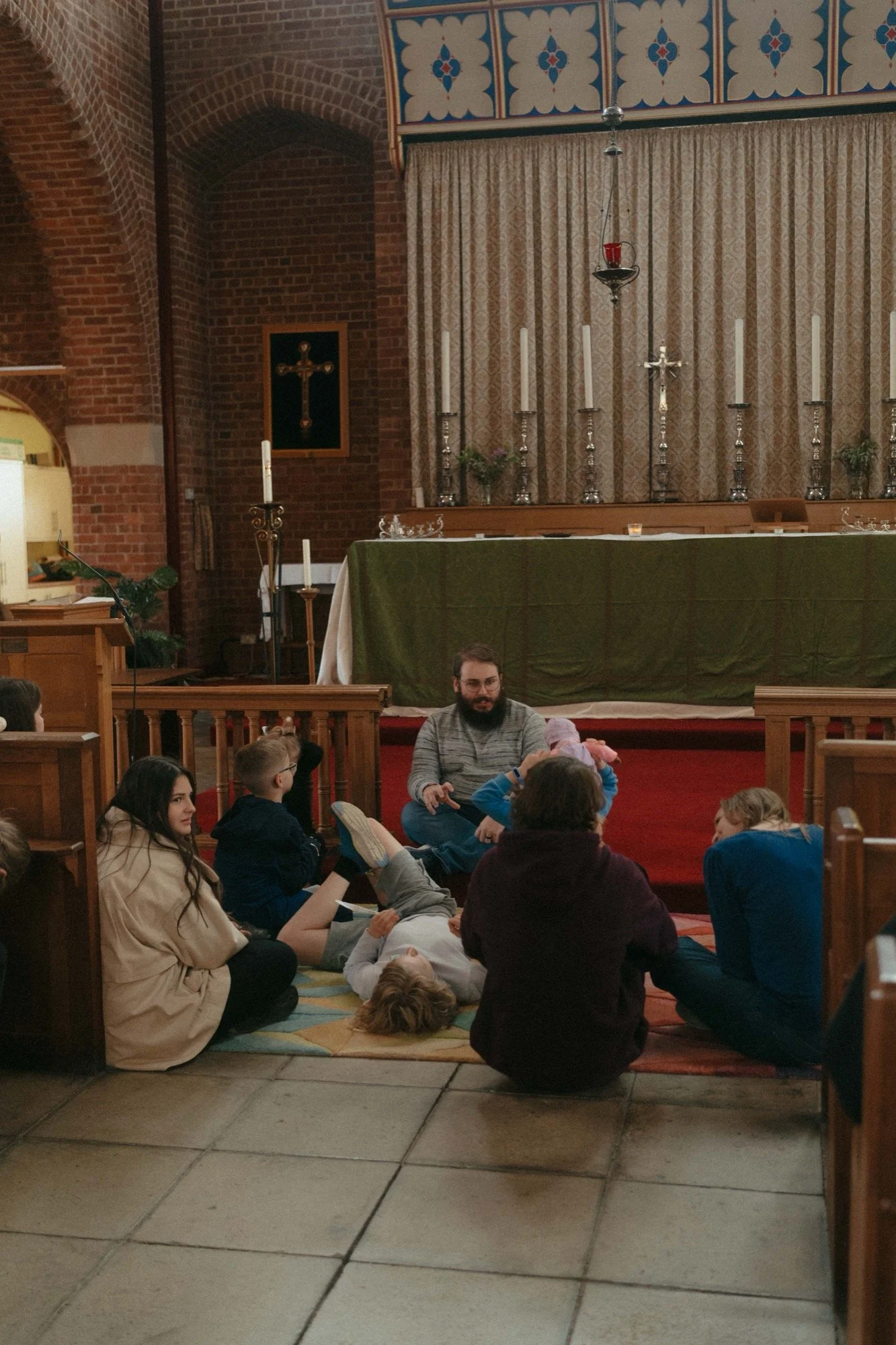 A man sits on the floor of a church, speaking to a group of children and a woman, with an altar and religious symbols in the background.