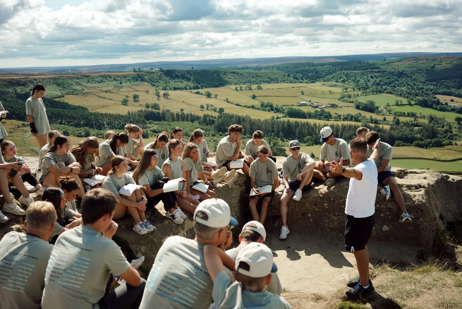 Group of young people sitting on rocks outdoors, listening to Mike Taylor speaking, with a scenic landscape of rolling green hills in the background.