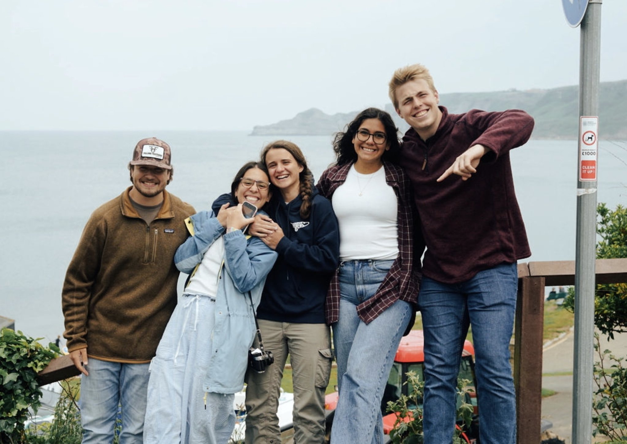 Group of five friends smiling and posing together outdoors near the coast, with the ocean and green hills in the background, during a cloudy day.