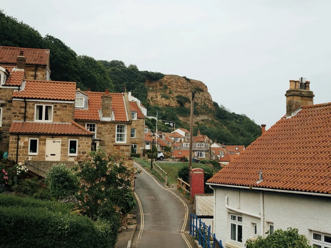 A small residential neighborhood with houses featuring red-tiled roofs, a narrow winding street, greenery, and a hillside with a rocky formation in the background.