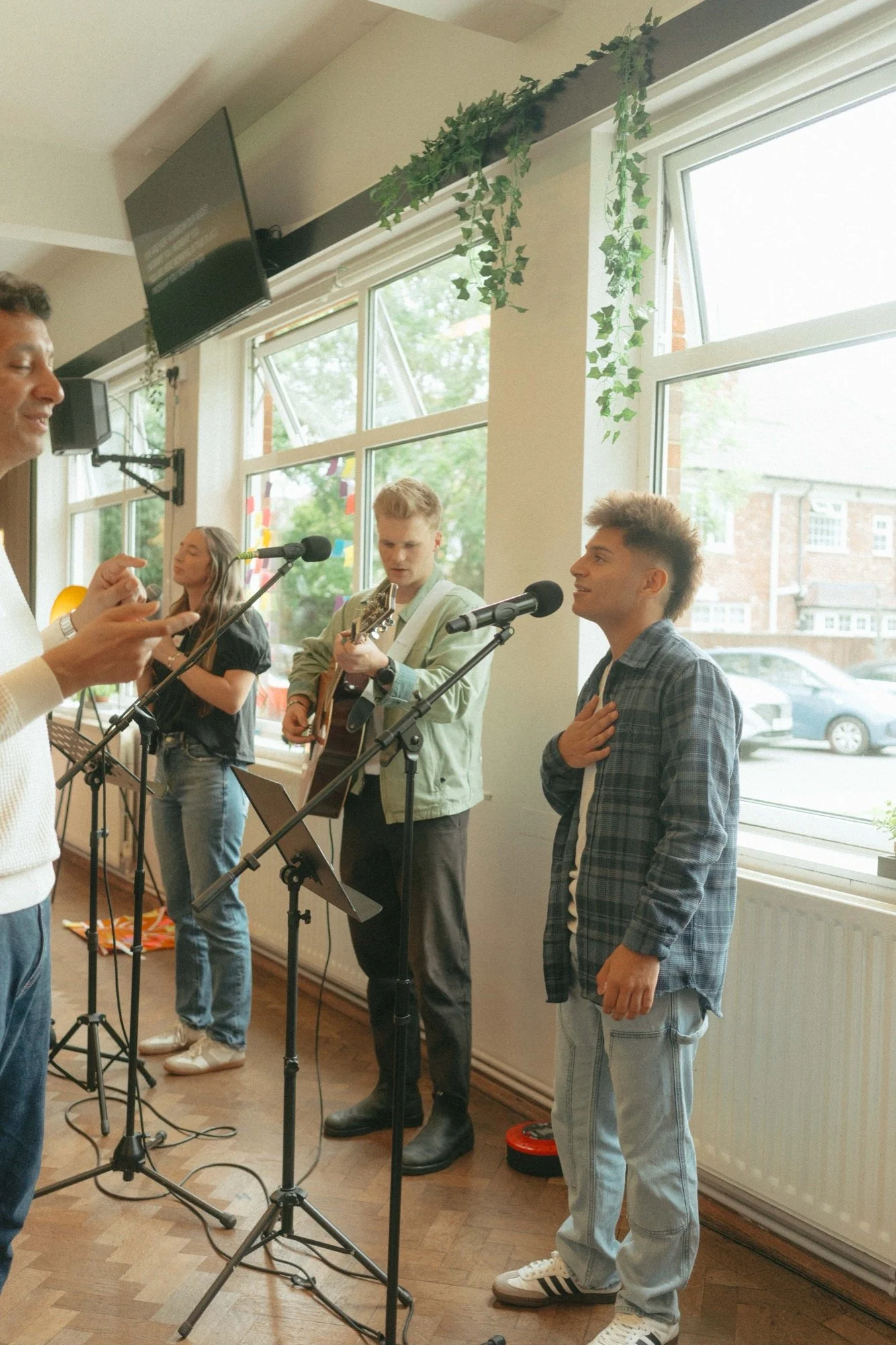 Group of young people performing music in a bright room with large windows, plants, and a television on the wall.