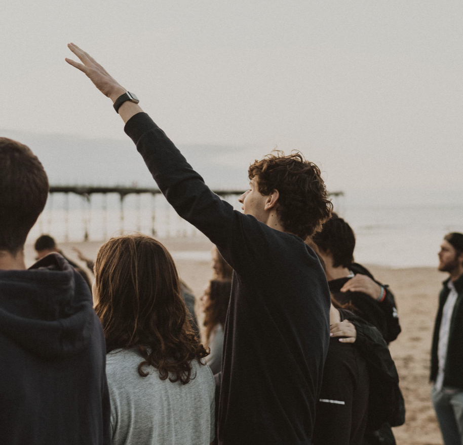 Group of young adults on a beach, with one person raising their hand, during sunset or early evening.