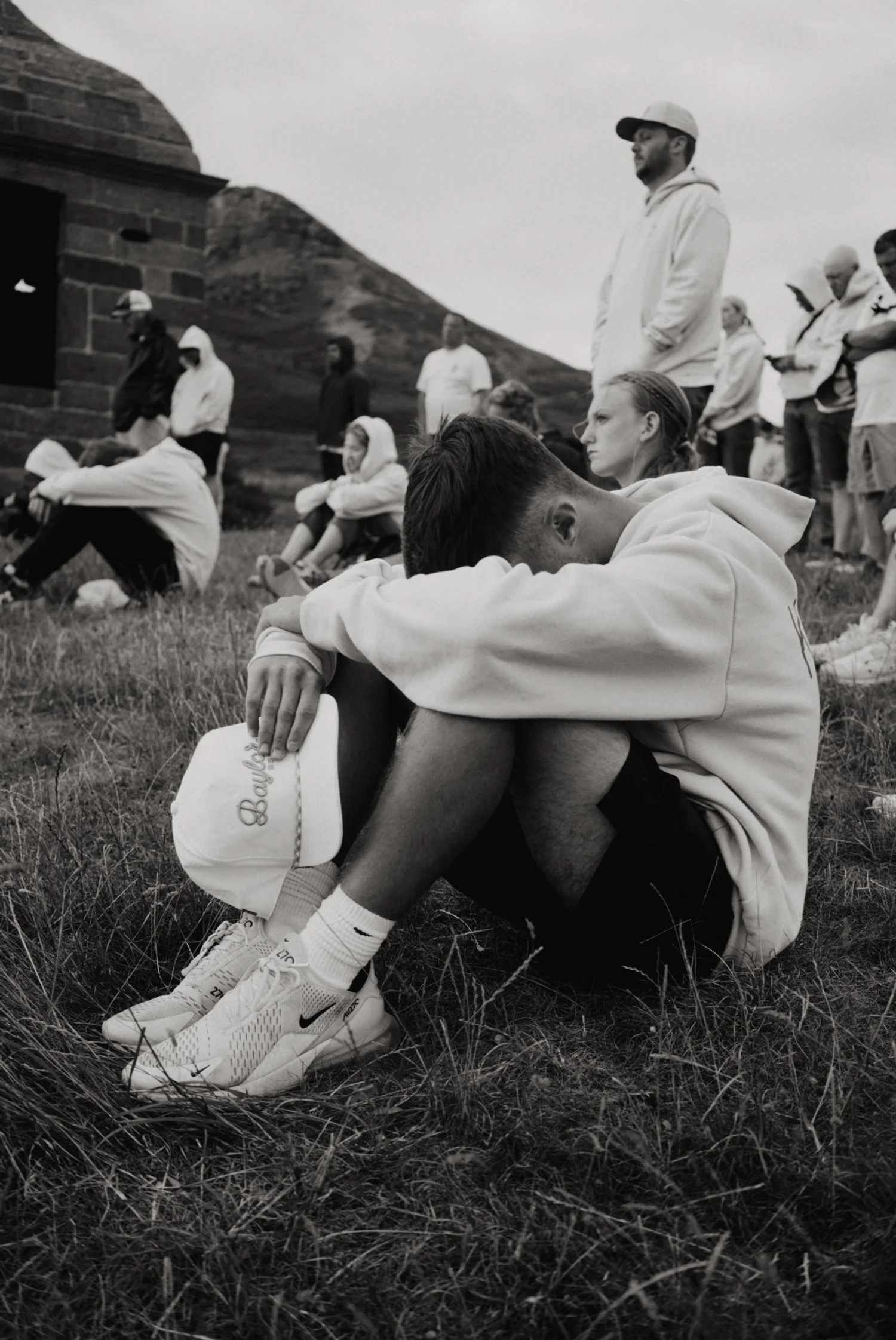 A young man sits on the grass with his head resting on his knees, holding a baseball cap. Other people are gathered around on a hillside, some sitting and some standing, with a hill and a stone building in the background.