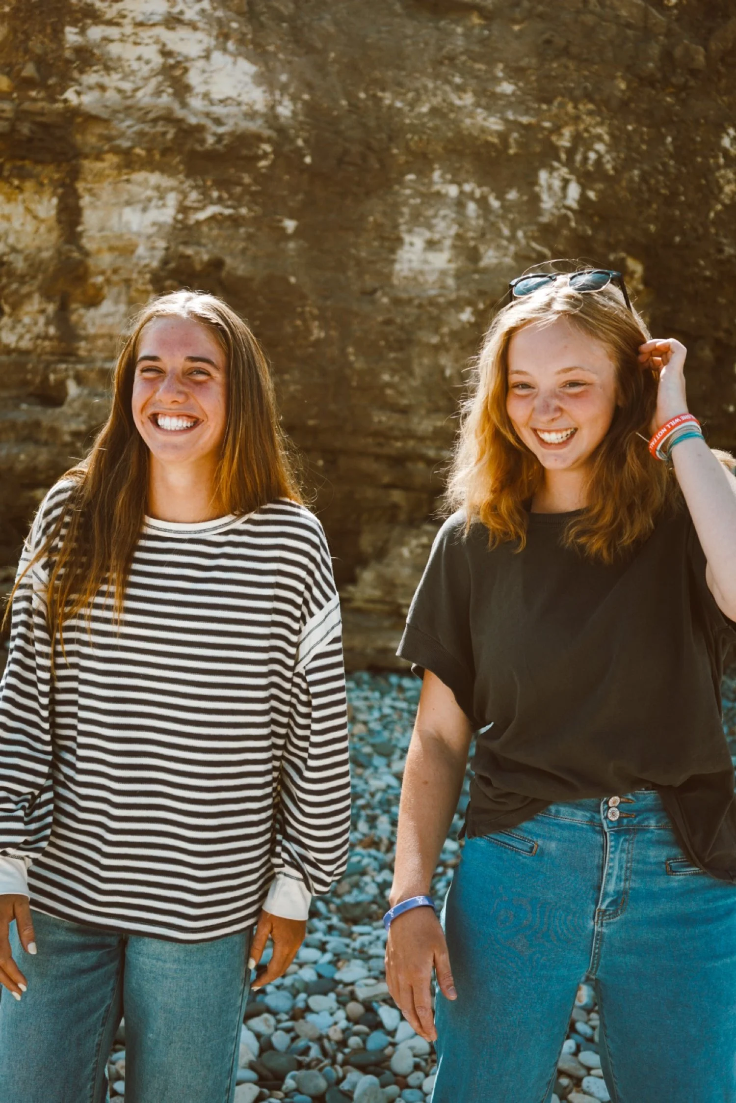Two young women smiling and laughing outdoors near a rocky cliff, with one wearing a black t-shirt and denim shorts and the other a striped long-sleeve shirt and jeans.