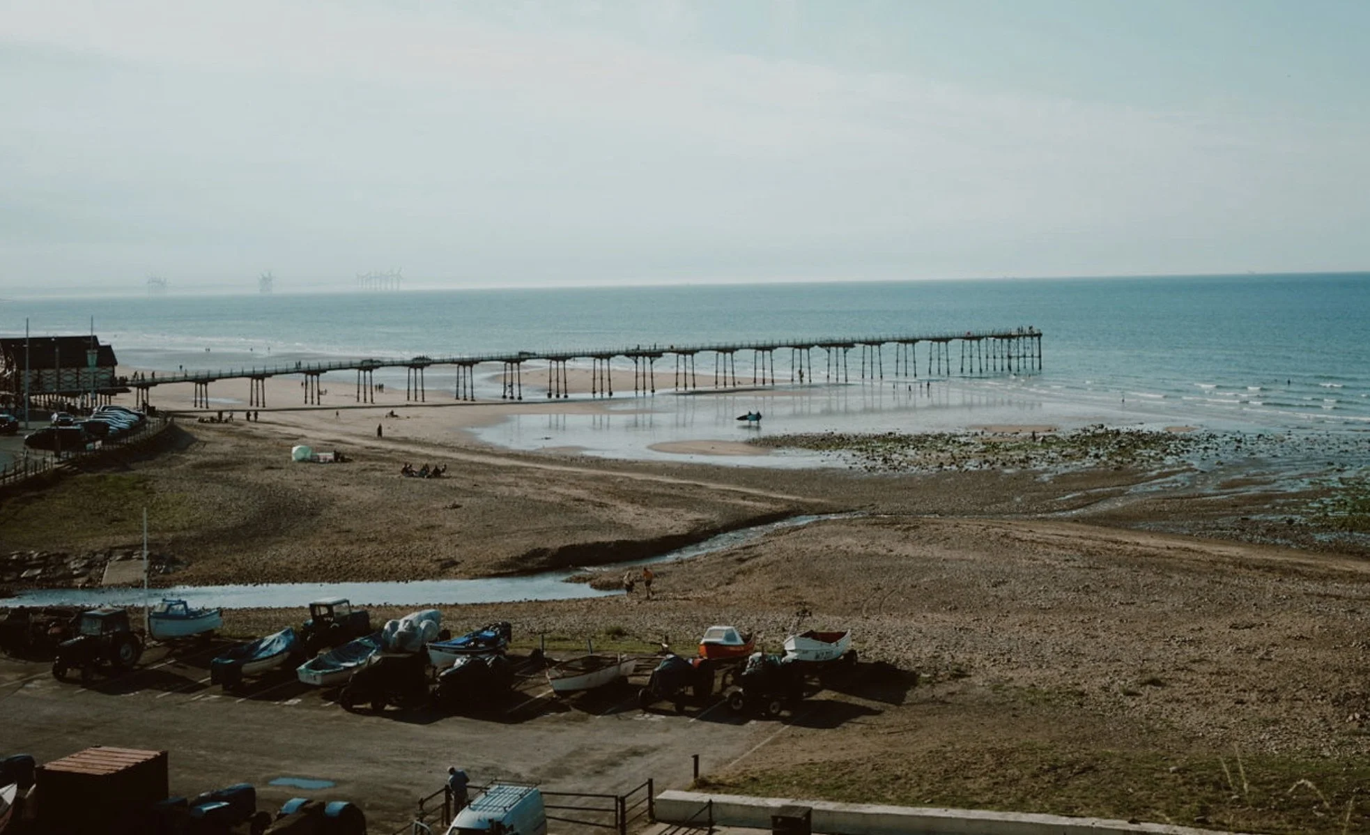 View of a beach with a long wooden pier extending into the ocean, boats on the sand, and parked cars and boats in the foreground. Small groups of people are visible on the beach and near the water, with an oil rig in the distance across the water.