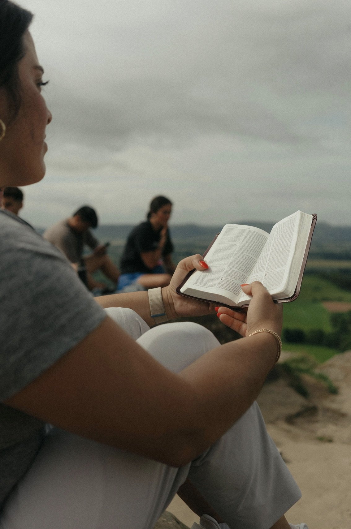 Woman reading a book outdoors with a rocky landscape and cloudy sky, accompanied by others sitting in the background.