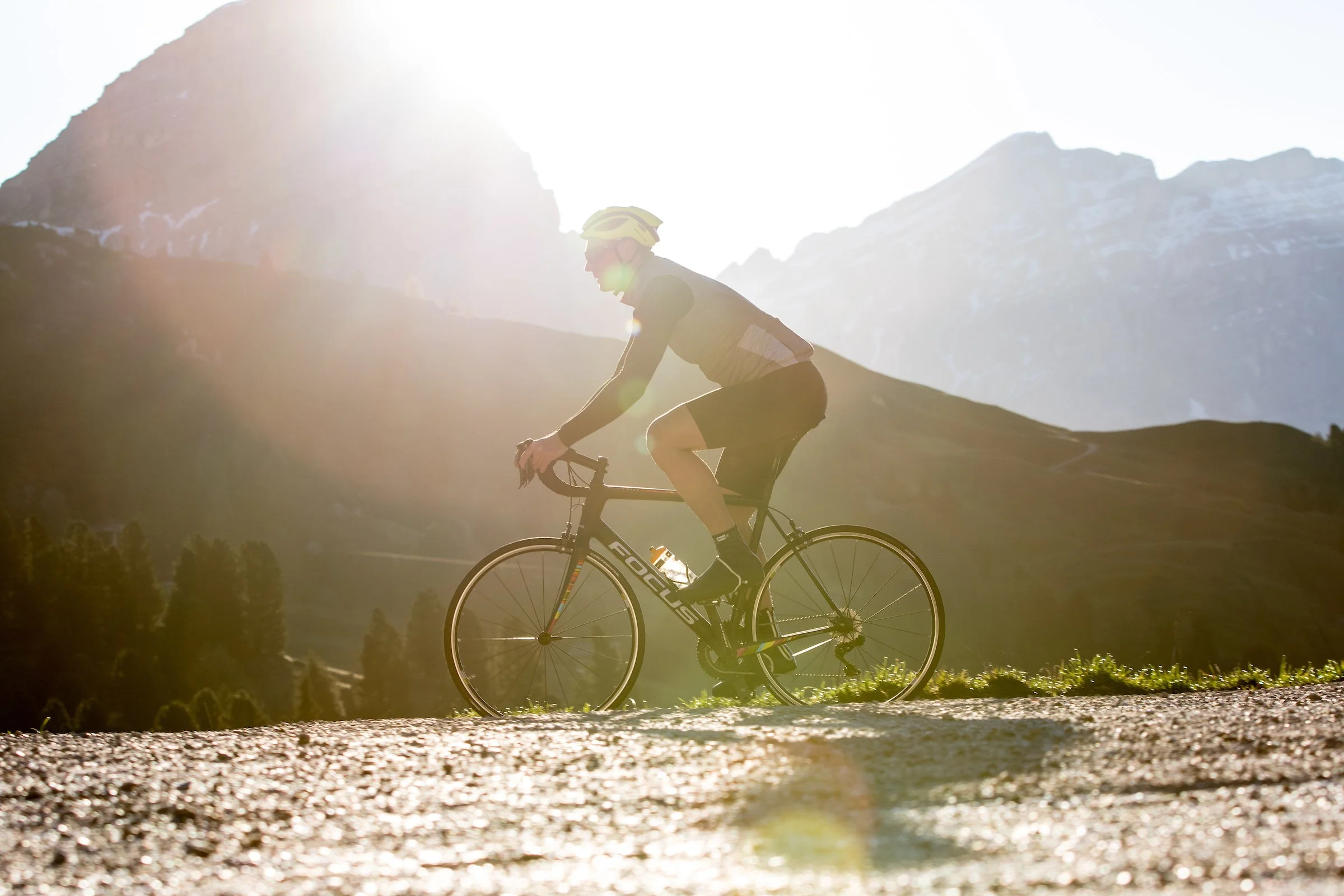 A person riding a bicycle outdoors with mountains in the background and sunlight creating a lens flare.