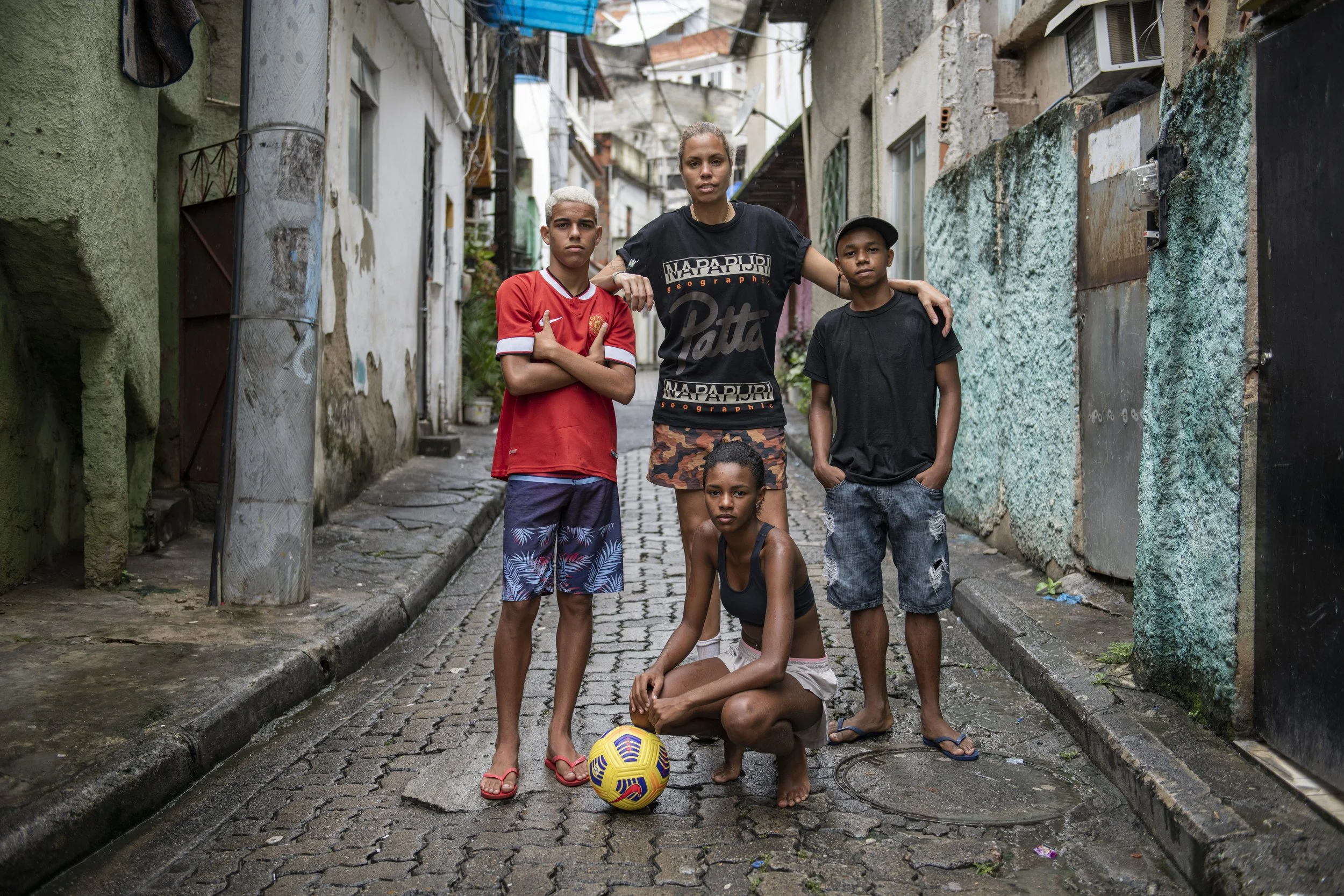 Group of five children and teenager in an alleyway with colorful, weathered walls. One girl is sitting on the ground with a soccer ball, and the other four stand around her. The girl standing in the center has her arm around the shoulders of one boy, while another boy with crossed arms stands to her left, and a boy with hands in pockets stands to her right. A girl in a black tank top and shorts is kneeling on the ground in front of them.