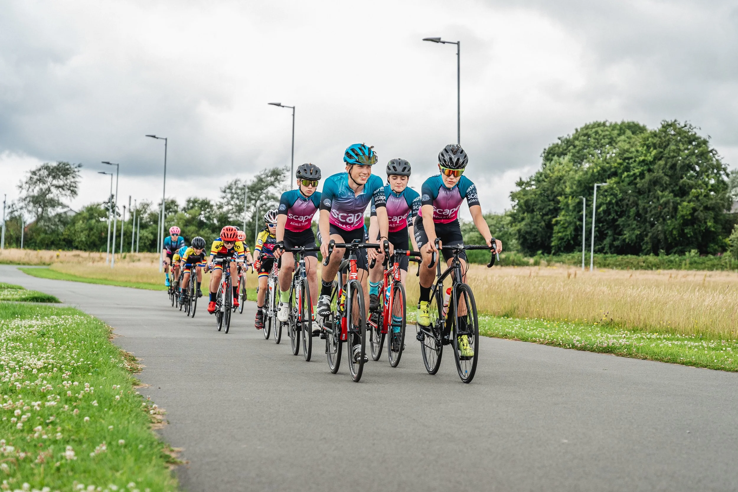 Cyclists participating in a group bike ride on a paved country road surrounded by green fields and trees, under a cloudy sky.