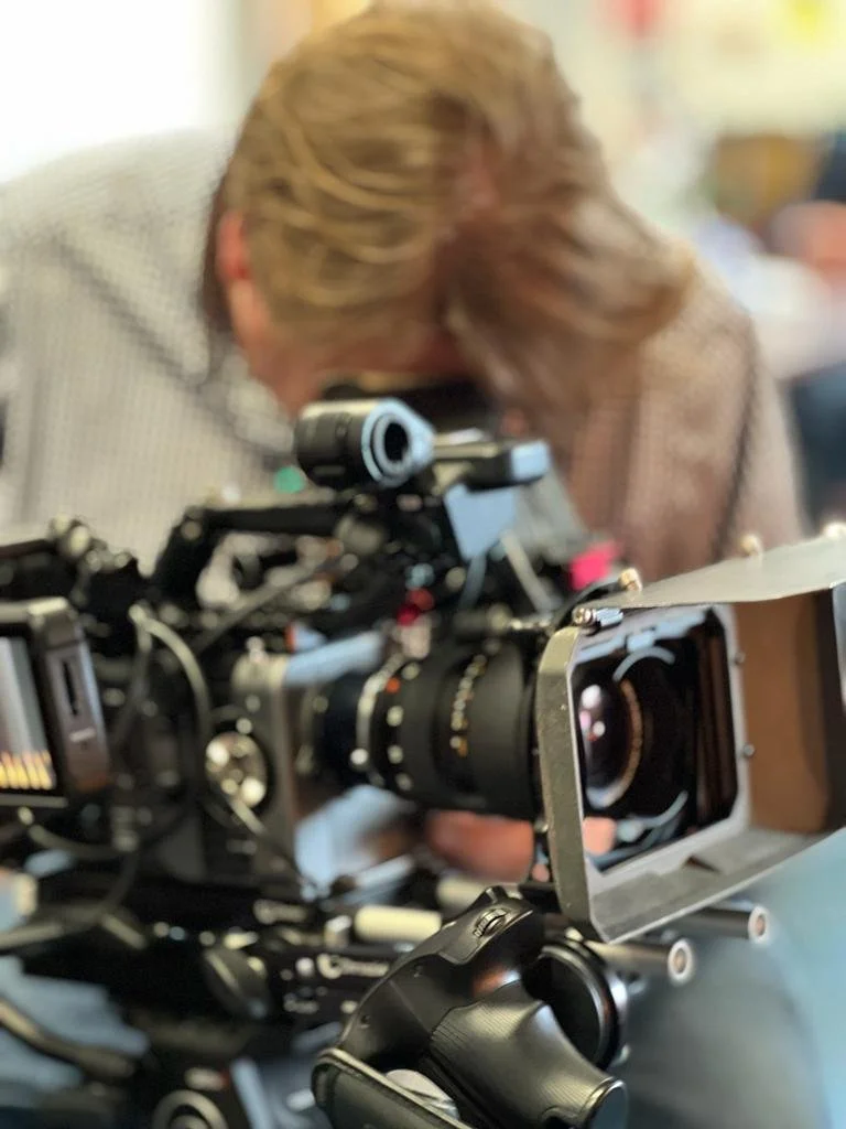 Close-up of professional film camera on set, with a woman in the background leaning in, possibly a director or crew member, partially blurred.