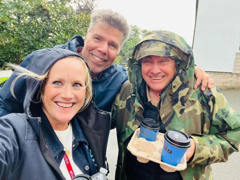 A group of three people smiling and taking a selfie outdoors. Two are women, and one is a man wearing military camouflage. The woman on the left has blonde hair and is wearing a dark jacket, the man on the right is dressed in camouflage with a hat, and the woman in the middle has short hair and is wearing a blue jacket. The man is holding two cups of coffee in a tray, and the background features green trees and a building.