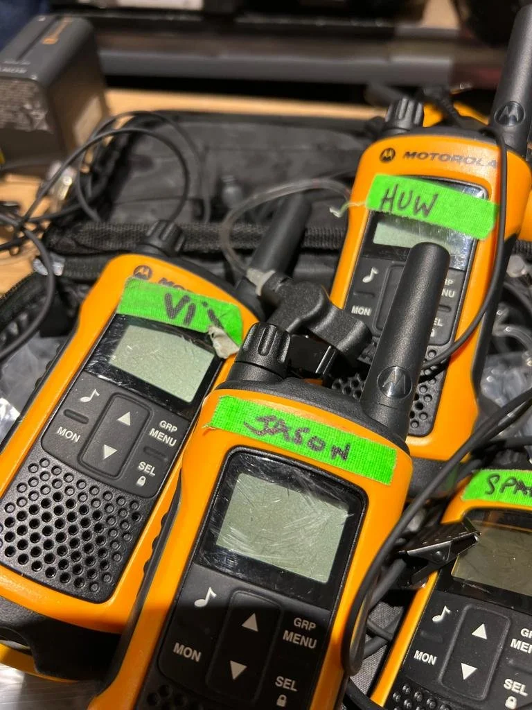 Four orange Motorola walkie-talkies with green labels reading 'VIA', 'JASON', 'HUW', and 'SPAR' are placed on a work surface with wires and other equipment around them.